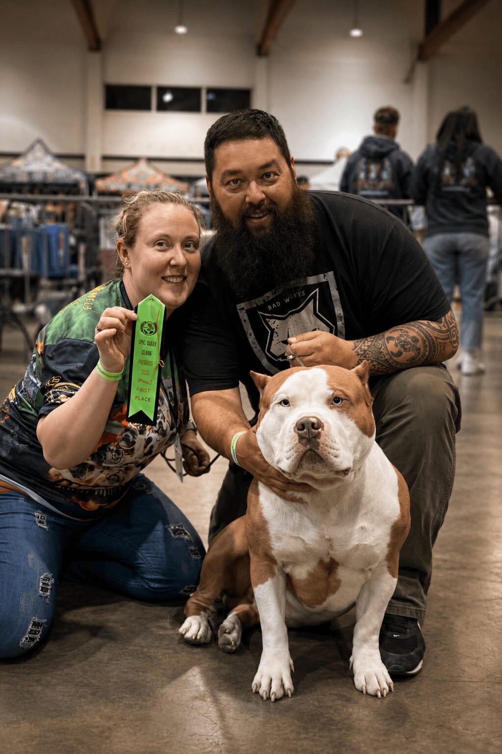 Two people, a woman and a man, pose with a large dog indoors at an event, smiling at the camera.
