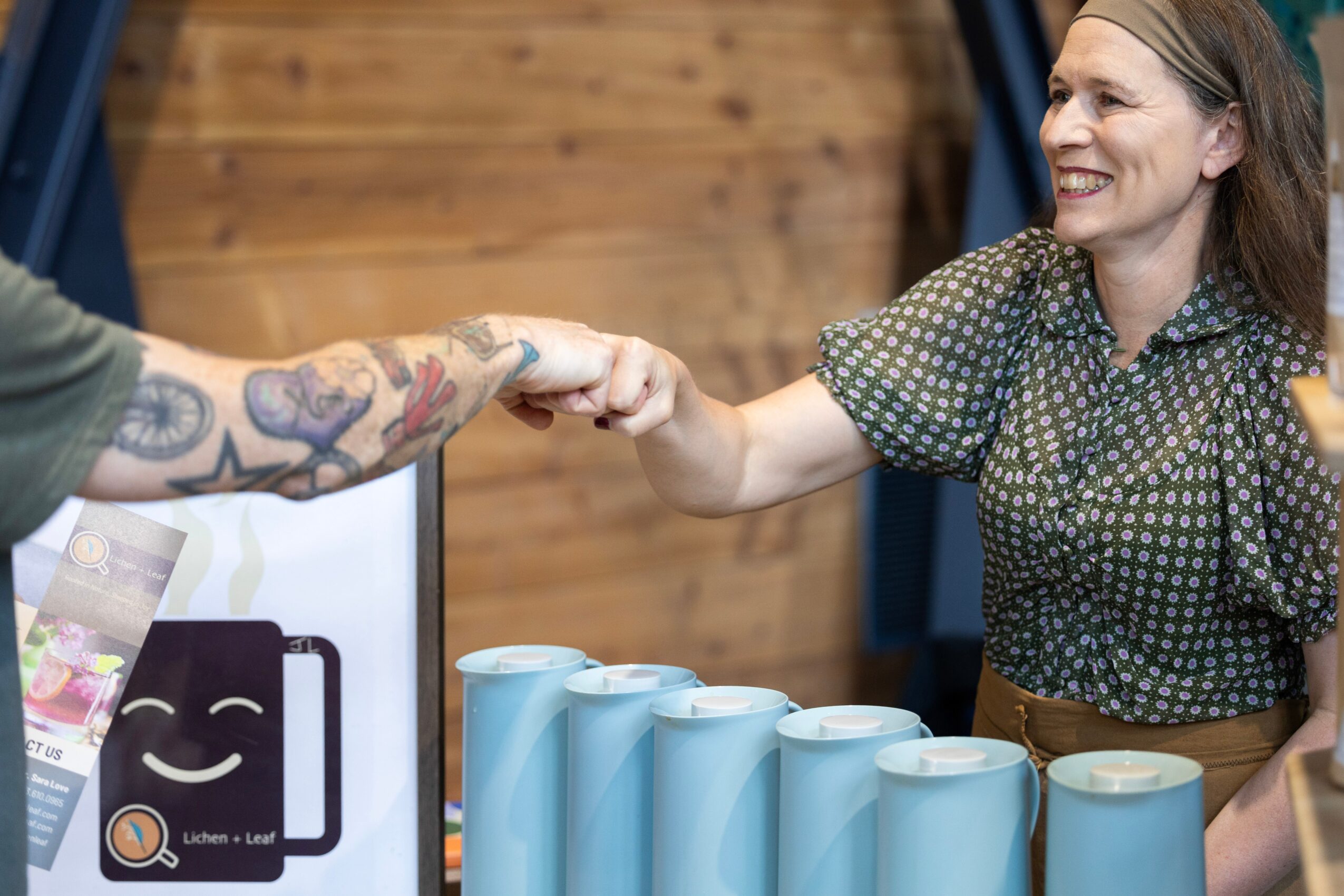 Two women shake hands in a room with wooden walls, smiling, with blue cups on the table.