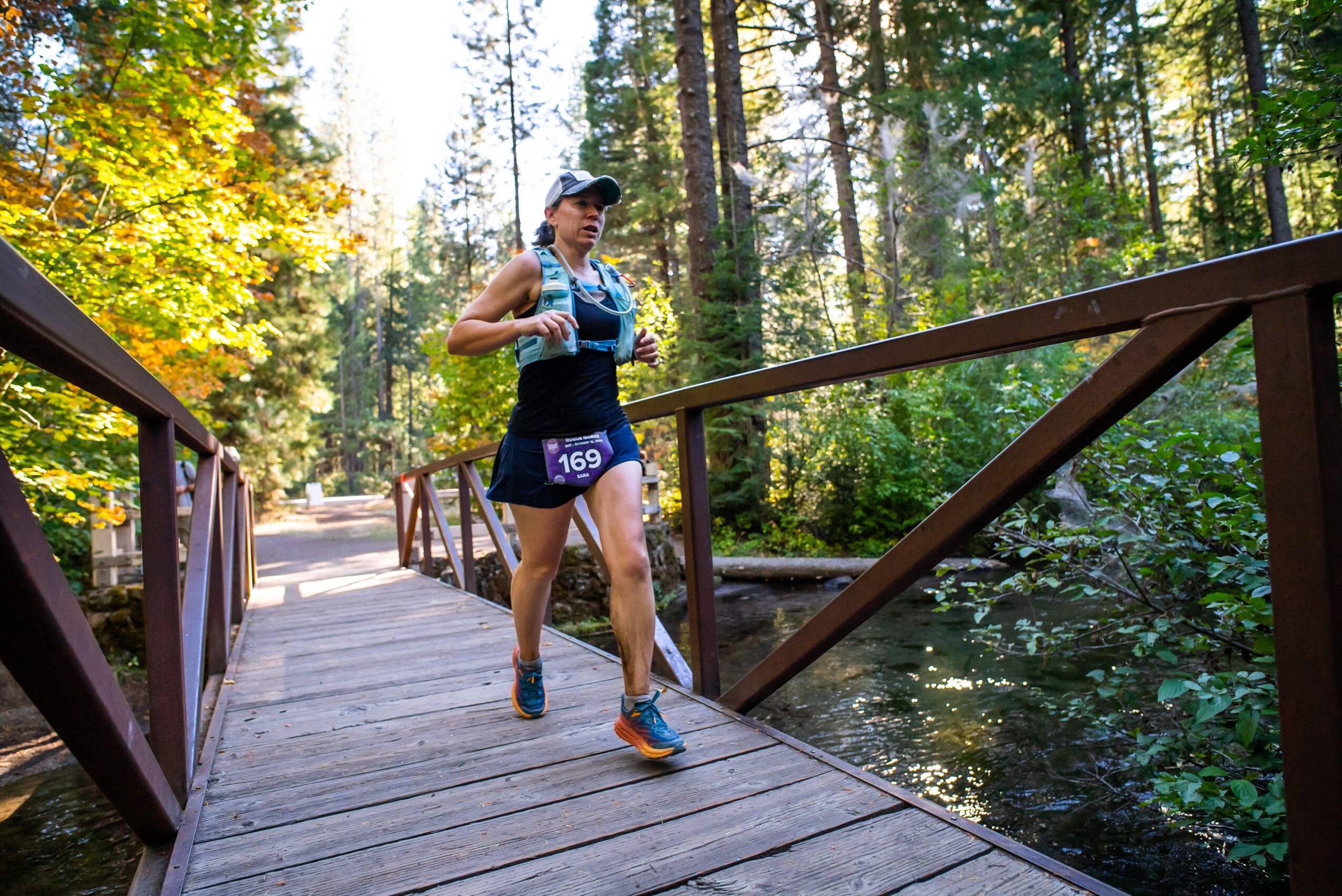 Woman running on a wooden bridge in a forest during daytime, wearing a cap, tank top, and shorts.