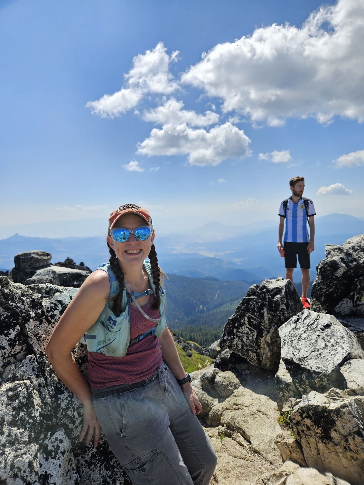 Two people on rocky mountain terrain under a blue sky with clouds, one sitting and one standing on rocks.