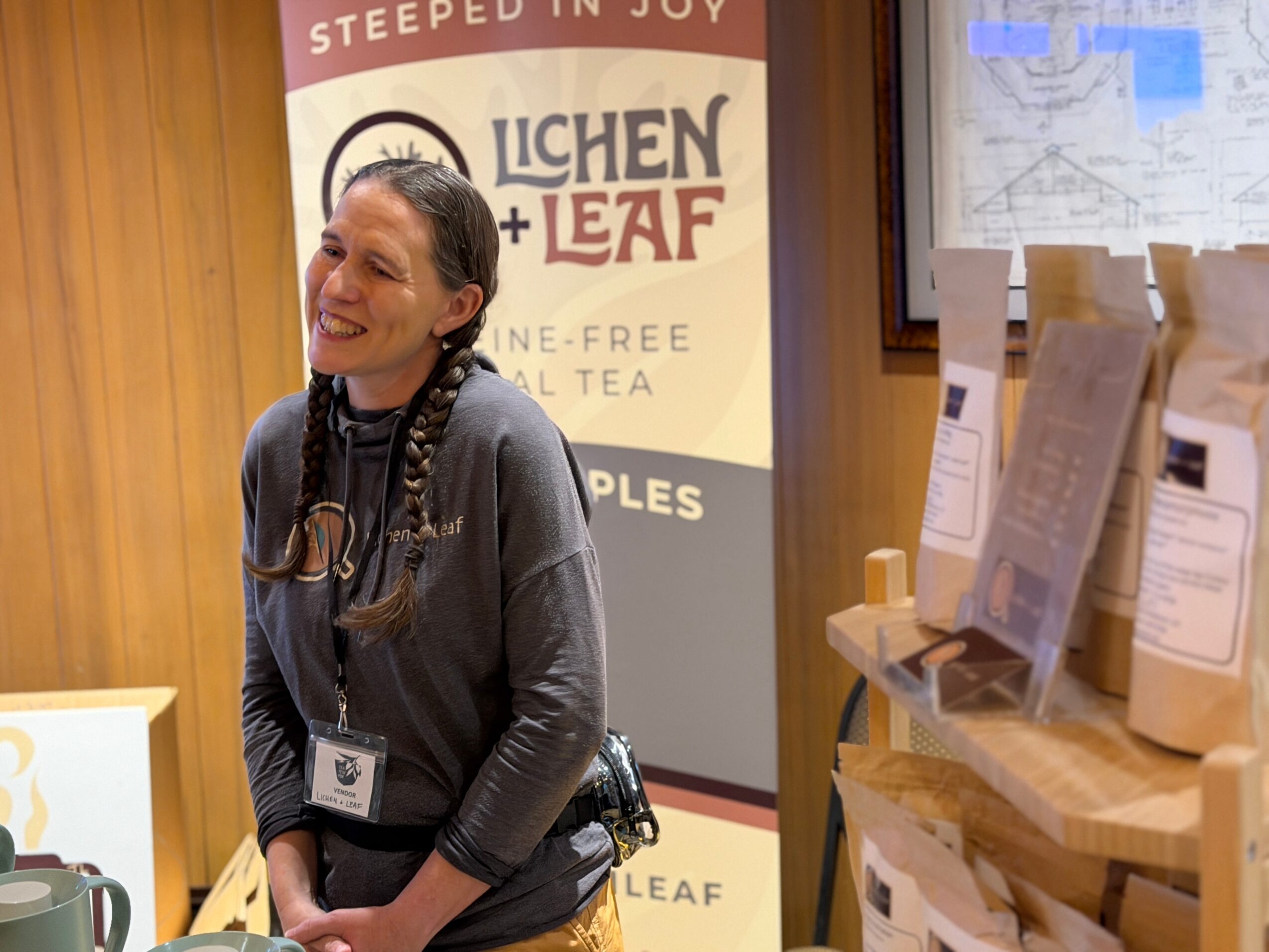 Smiling woman with braided hair and a badge stands in front of a tea display and banner, indoors.
