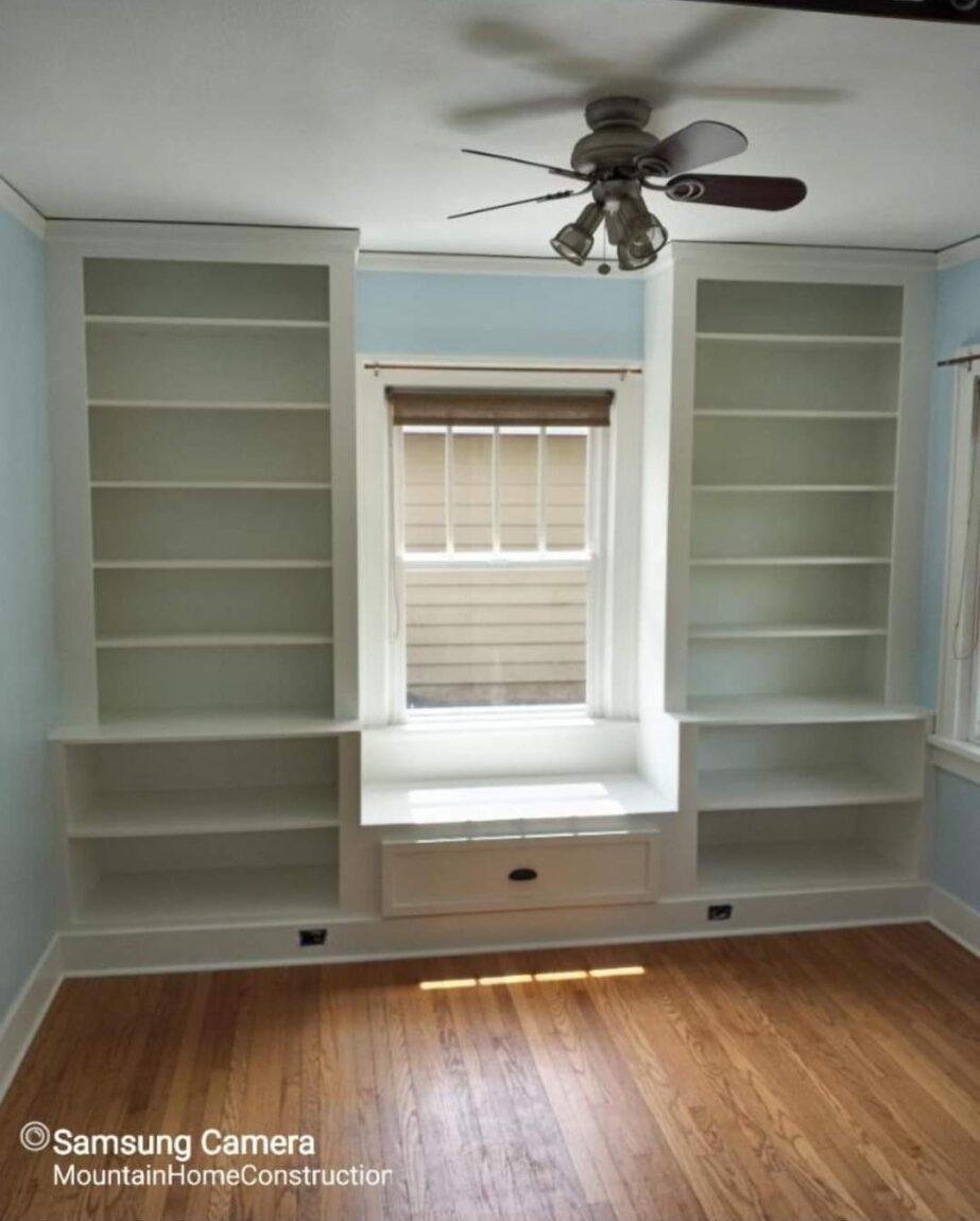 Room with built-in shelves on both sides of a window, ceiling fan, hardwood floor, and blinds, empty and well-lit.