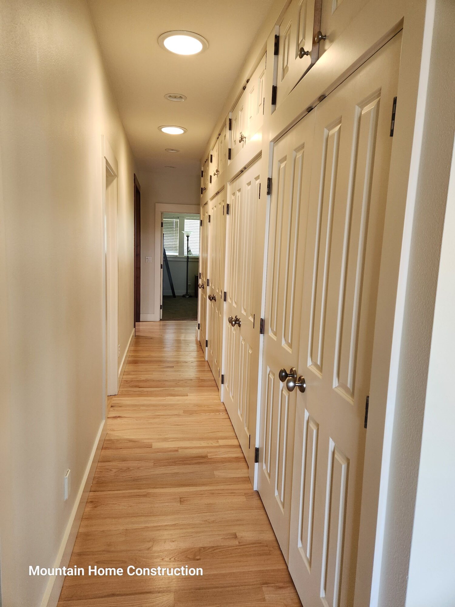 Long hallway with wooden flooring, white walls, ceiling lights, and a door at the end.