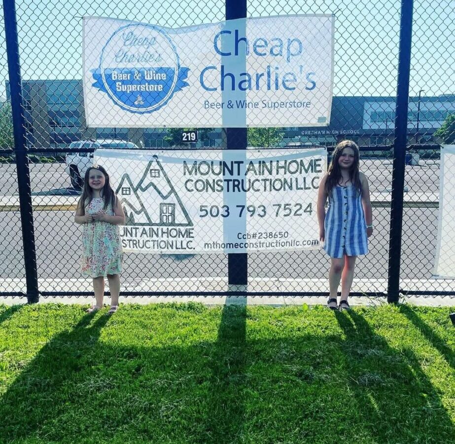 Two children stand on grass in front of a chain-link fence with a sign, under a dark sky.