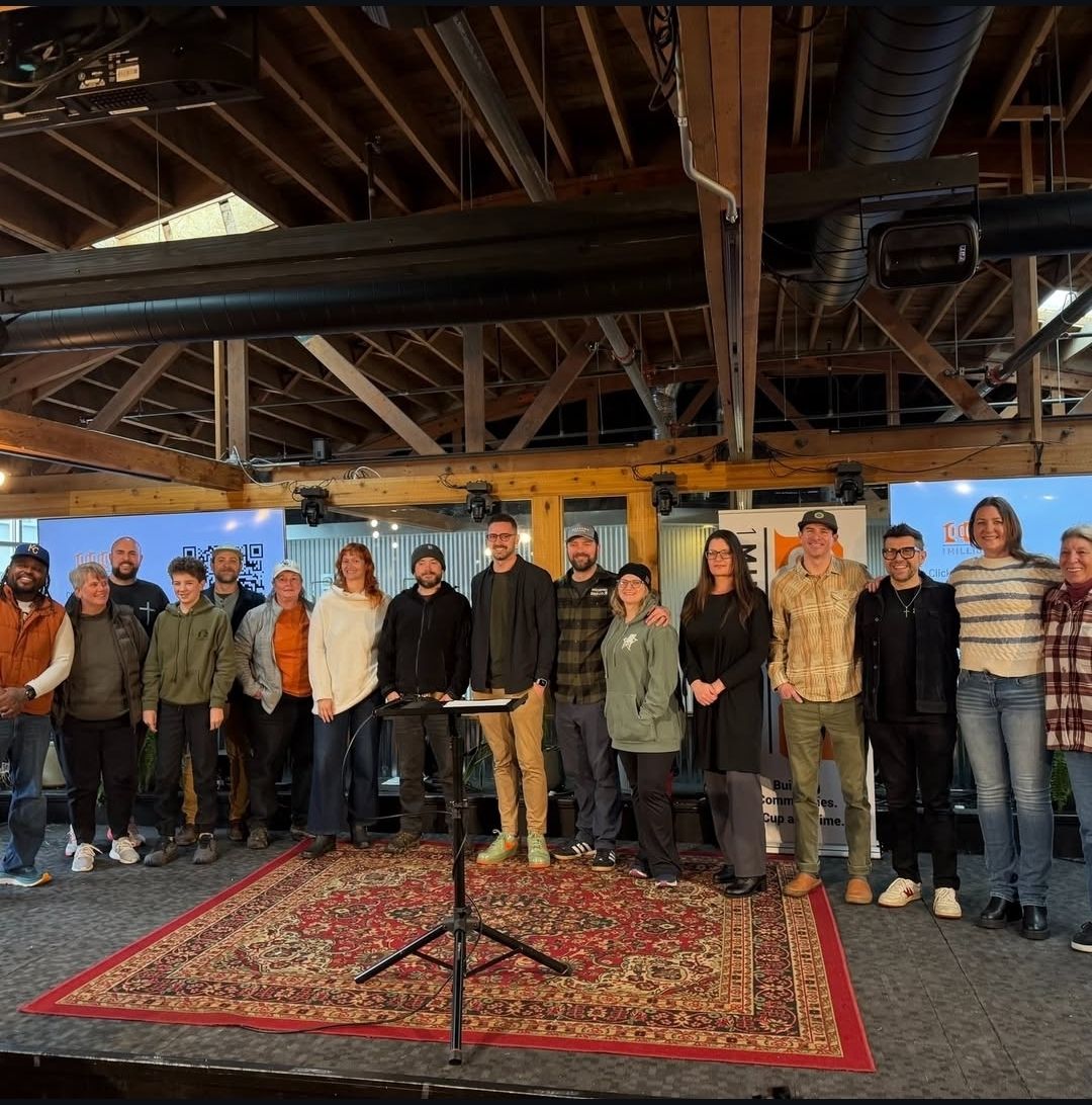 Group of people standing on a rug in an indoor space with exposed ceiling beams and pipes.