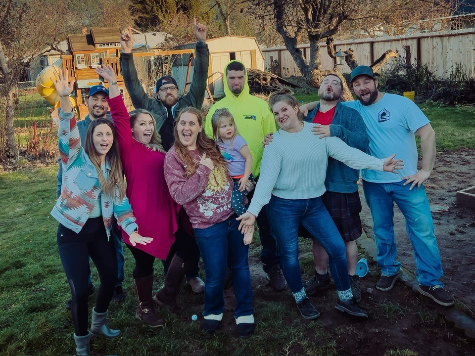 Group of ten people outdoors, smiling and posing, some with arms raised, in a yard with trees and a vehicle in background.