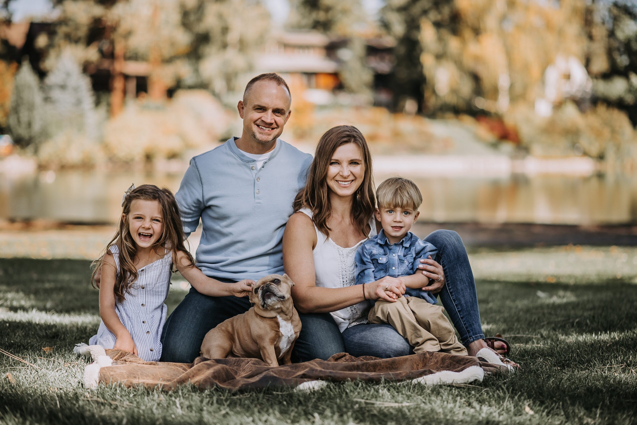Family of four with a dog sitting on a blanket outdoors near a lake, smiling, with trees and houses in background.