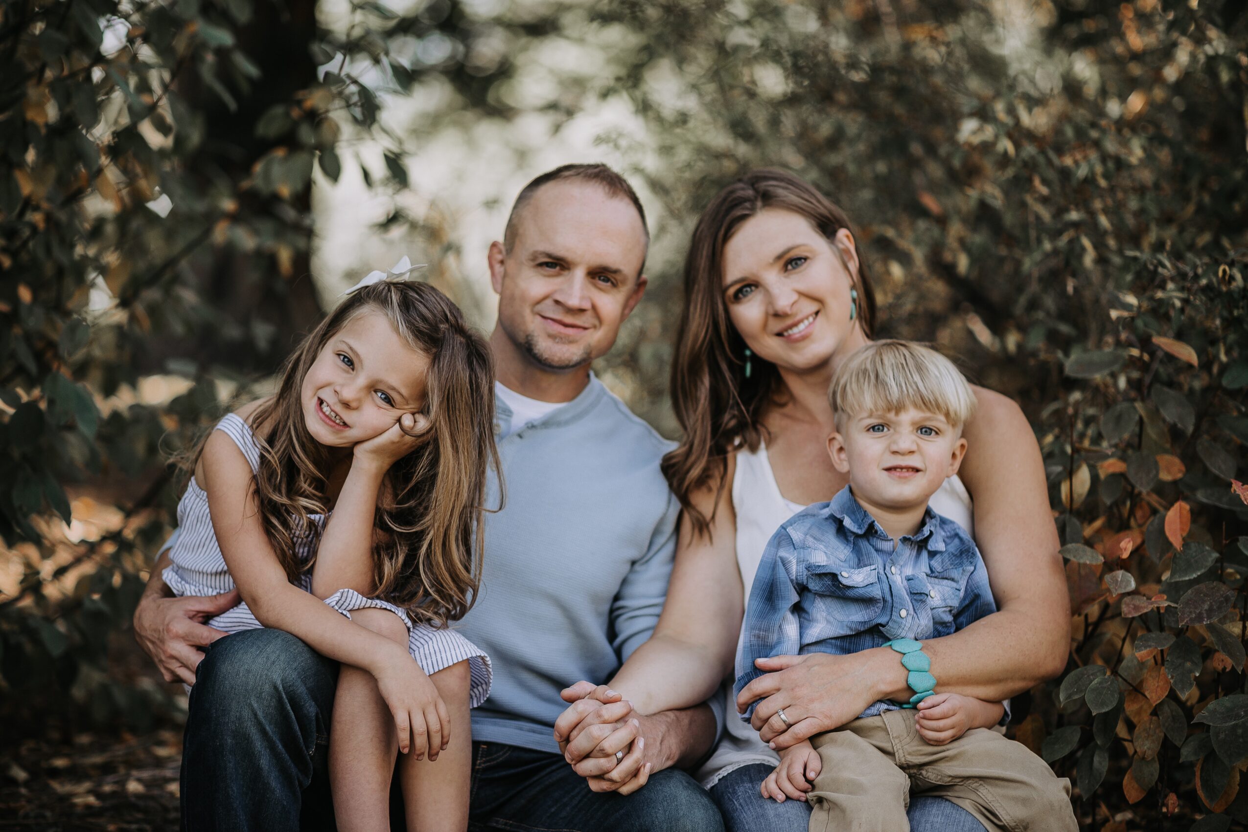 Family of four outdoors, sitting close together, smiling, surrounded by trees and foliage.