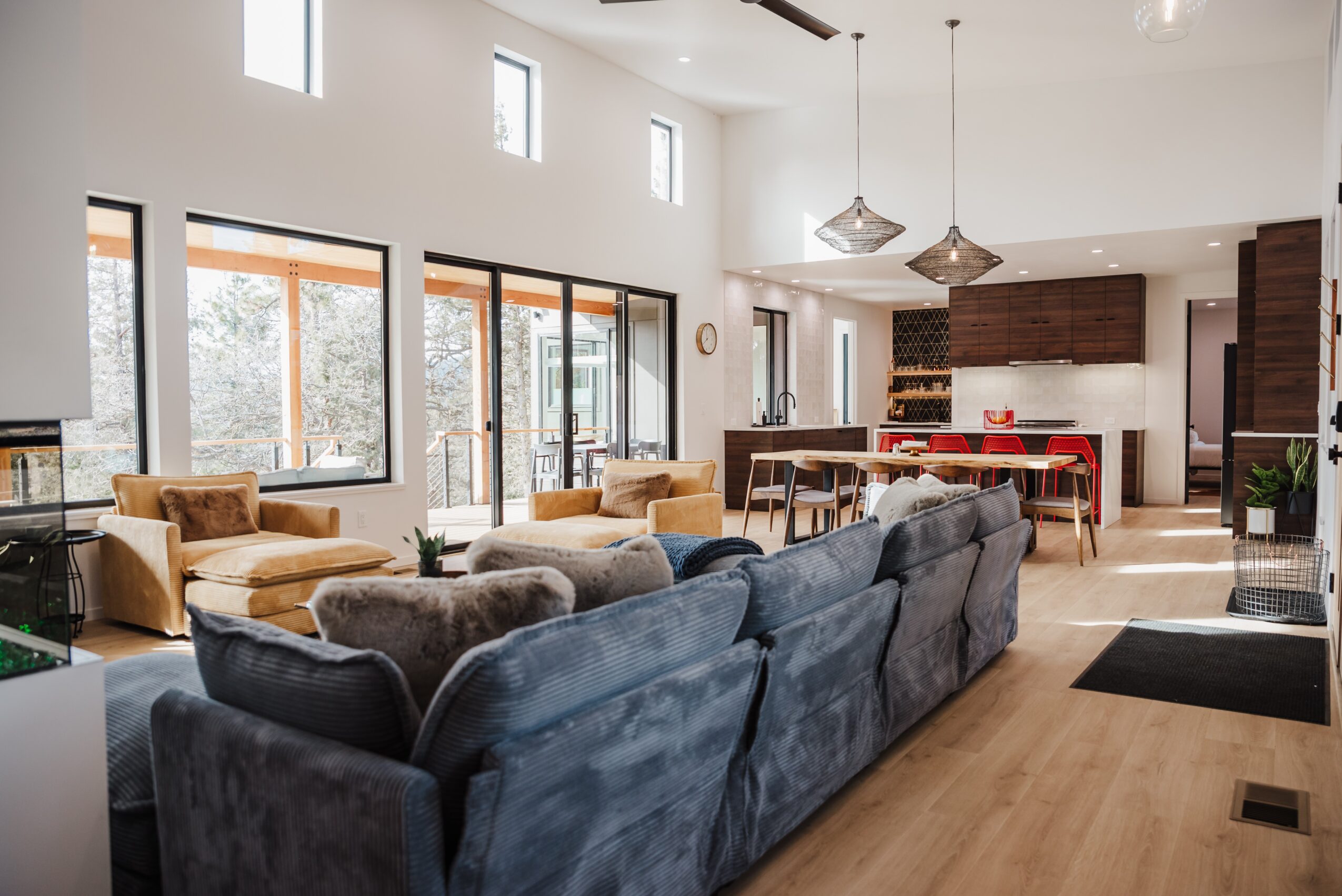 Spacious living room with large windows, sofas, and a view of the kitchen area with dining table, pendant lights, and cabinets.