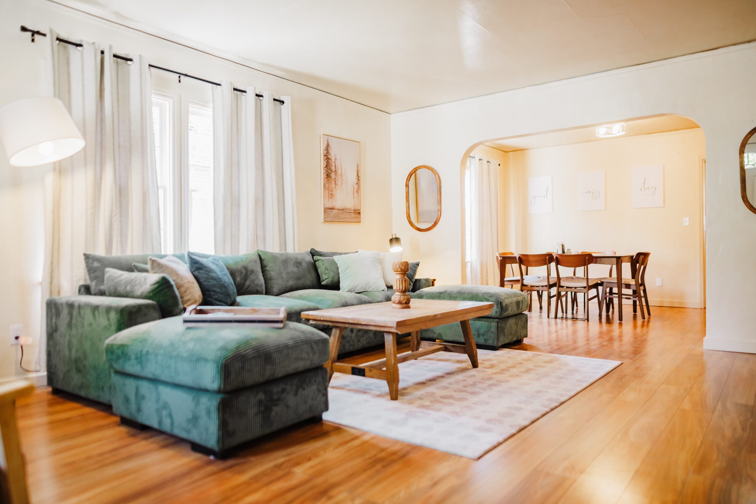 Living room with green sectional sofa, wooden coffee table, and dining area in background, bright natural light, hardwood floor.