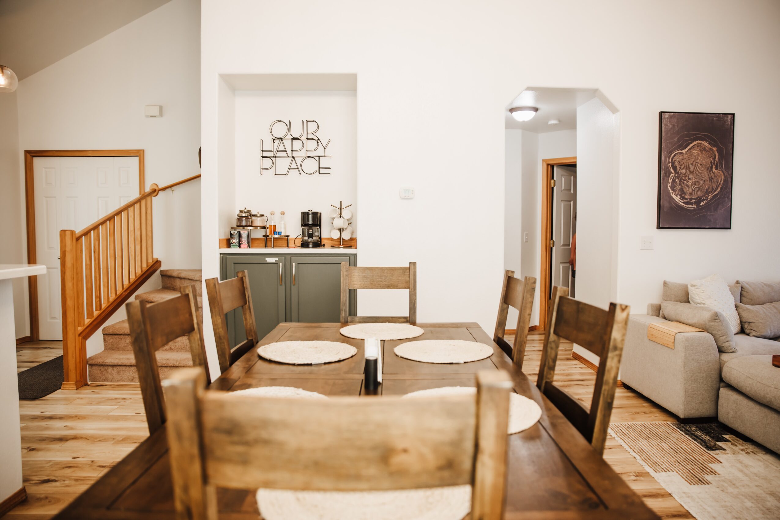 Dining area with wooden table and chairs, a small kitchen counter, and a sofa in the background, with artwork on the wall.