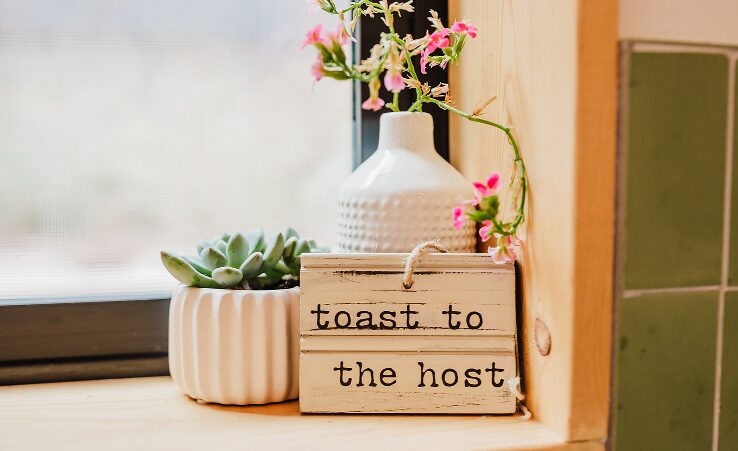 Decorative arrangement with a white vase holding pink flowers, a small potted succulent, and a wooden sign reading 'toast to the host' on a windowsill.