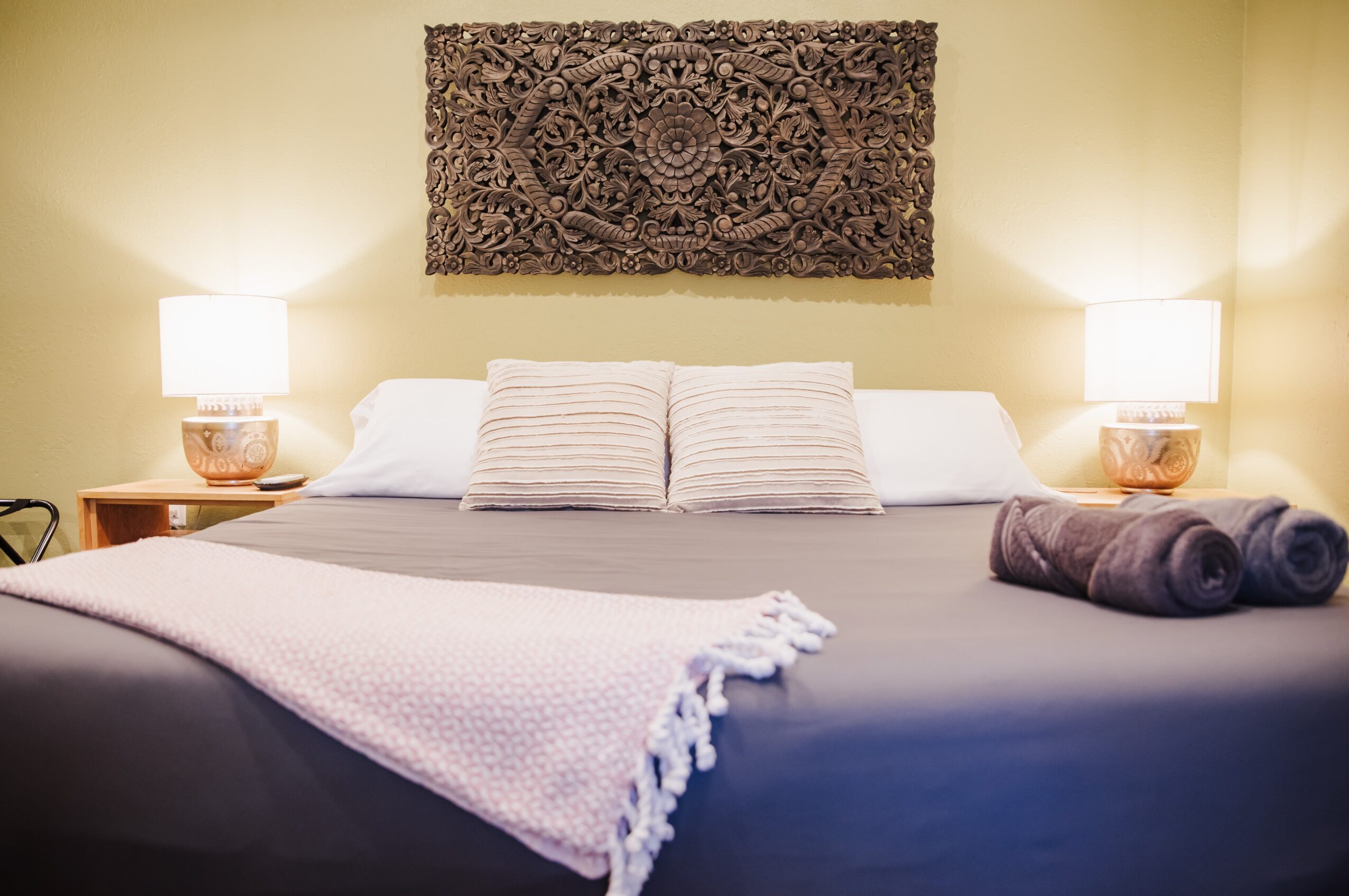 A neatly made bed with pillows, a blanket, and two rolled towels, flanked by two lamps on bedside tables, with wall art above.