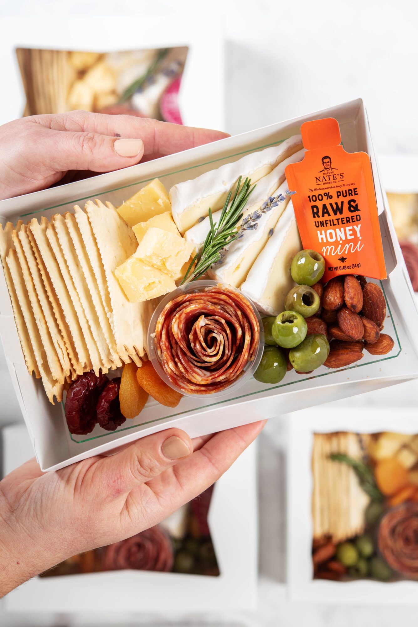 Hand holding a tray with cheese, crackers, green olives, nuts, honey, and a rose-shaped food item.