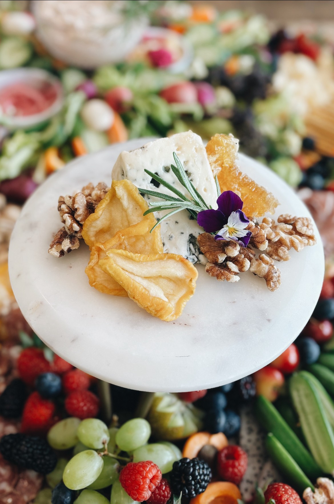 Cheesecake topped with walnuts, a yellow flower, and a purple flower, on a white plate with fruit and flowers in background.