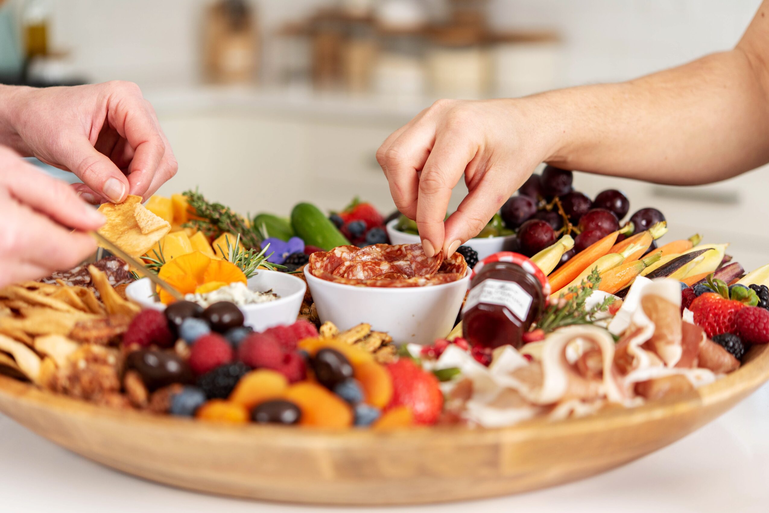 People's hands reaching for food on a large platter with fruits, cheese, and condiments, in a bright kitchen setting.