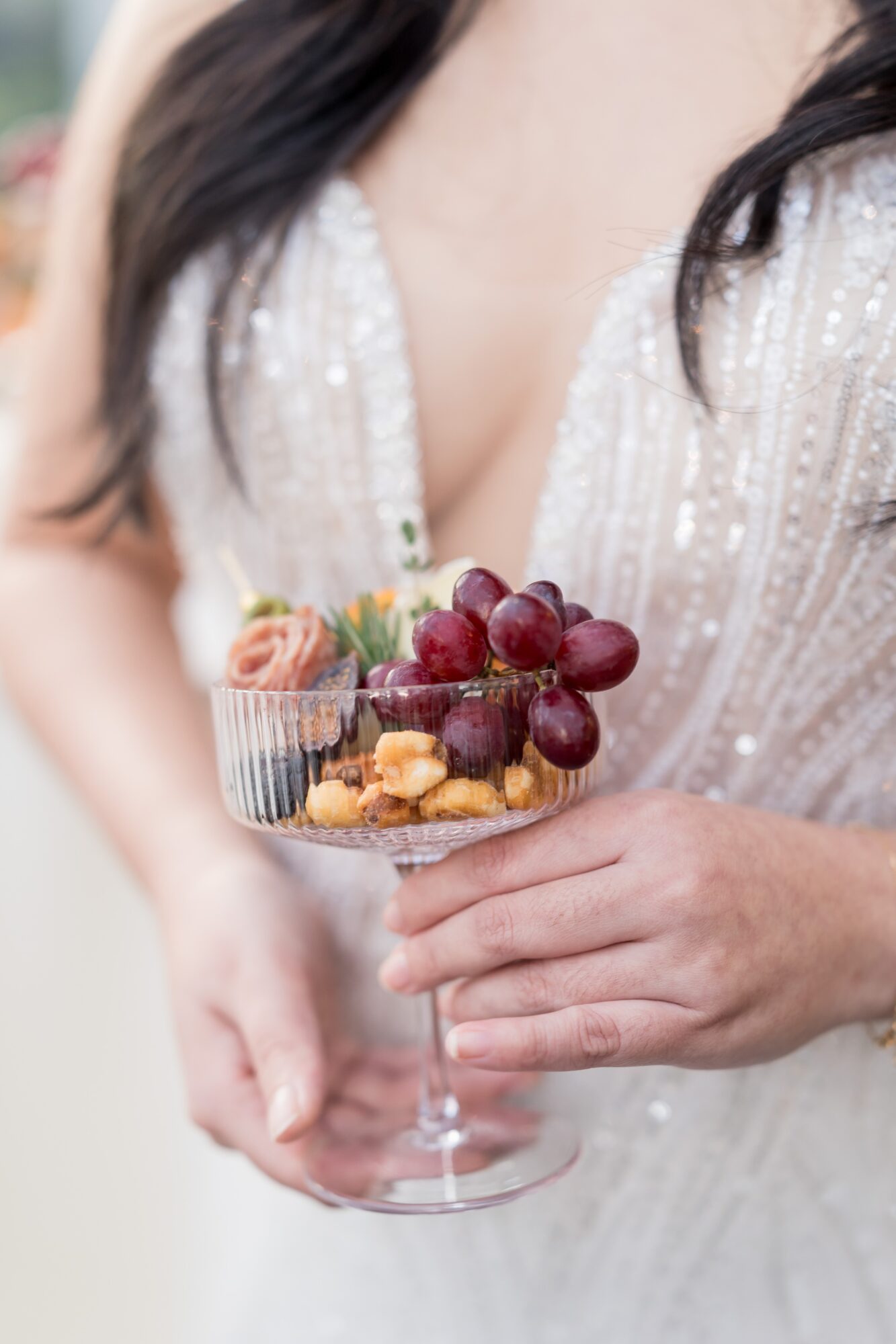 Person holding a glass bowl with grapes, cookies, and strawberries, wearing a white, beaded dress.