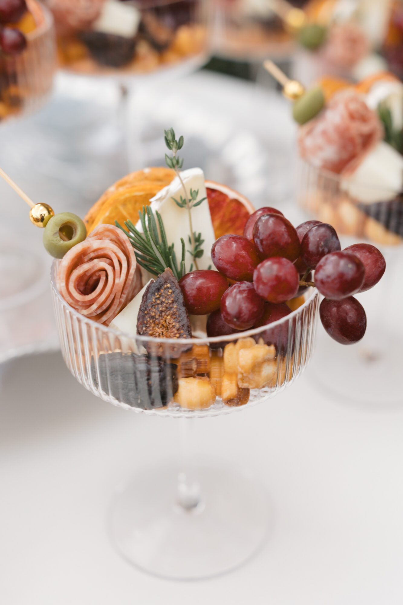 Glass dessert bowl with grapes, cheese, and fruit garnishes, on a table with blurred desserts in background.
