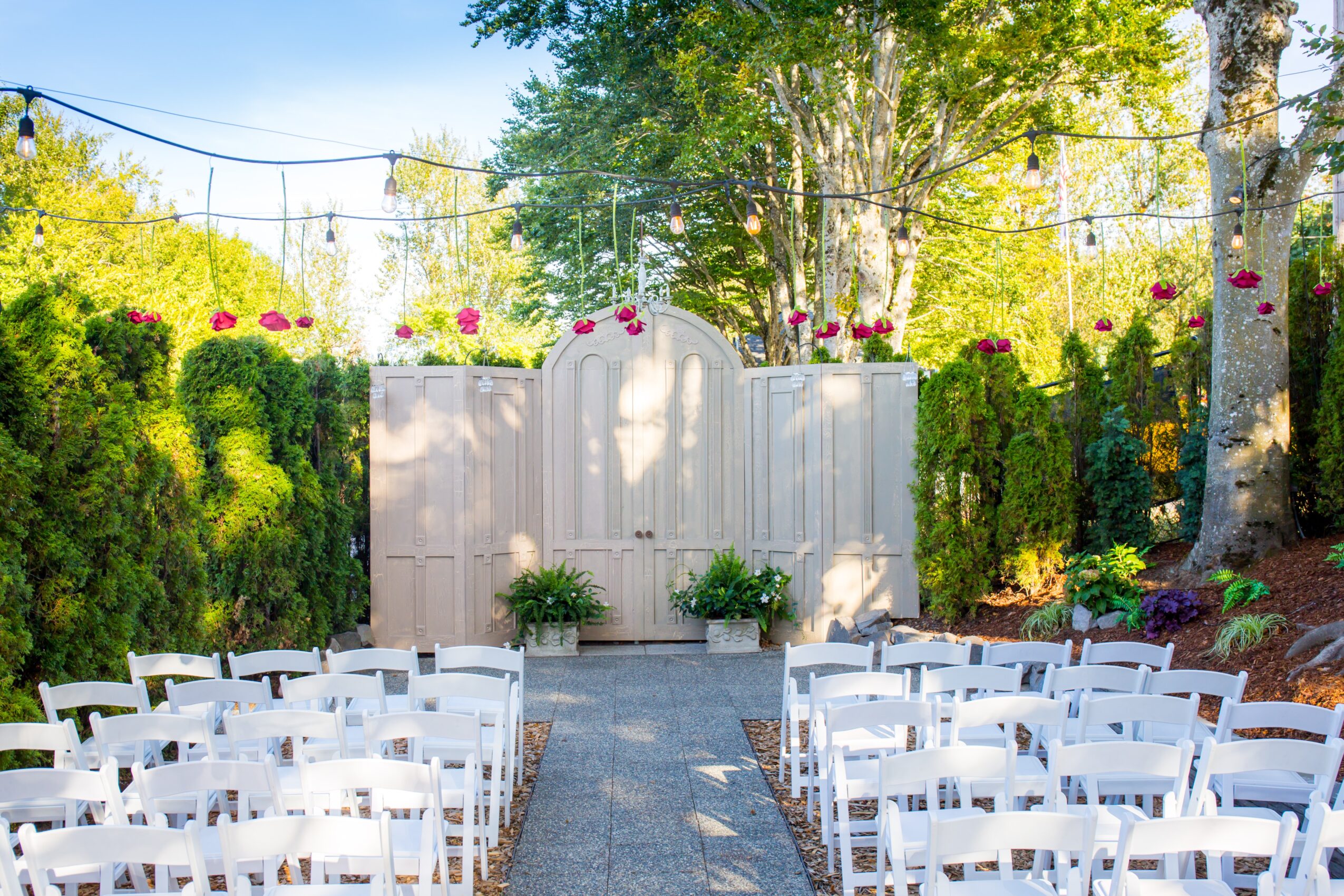 Outdoor wedding ceremony setup with white chairs facing a decorated backdrop, surrounded by trees and greenery.