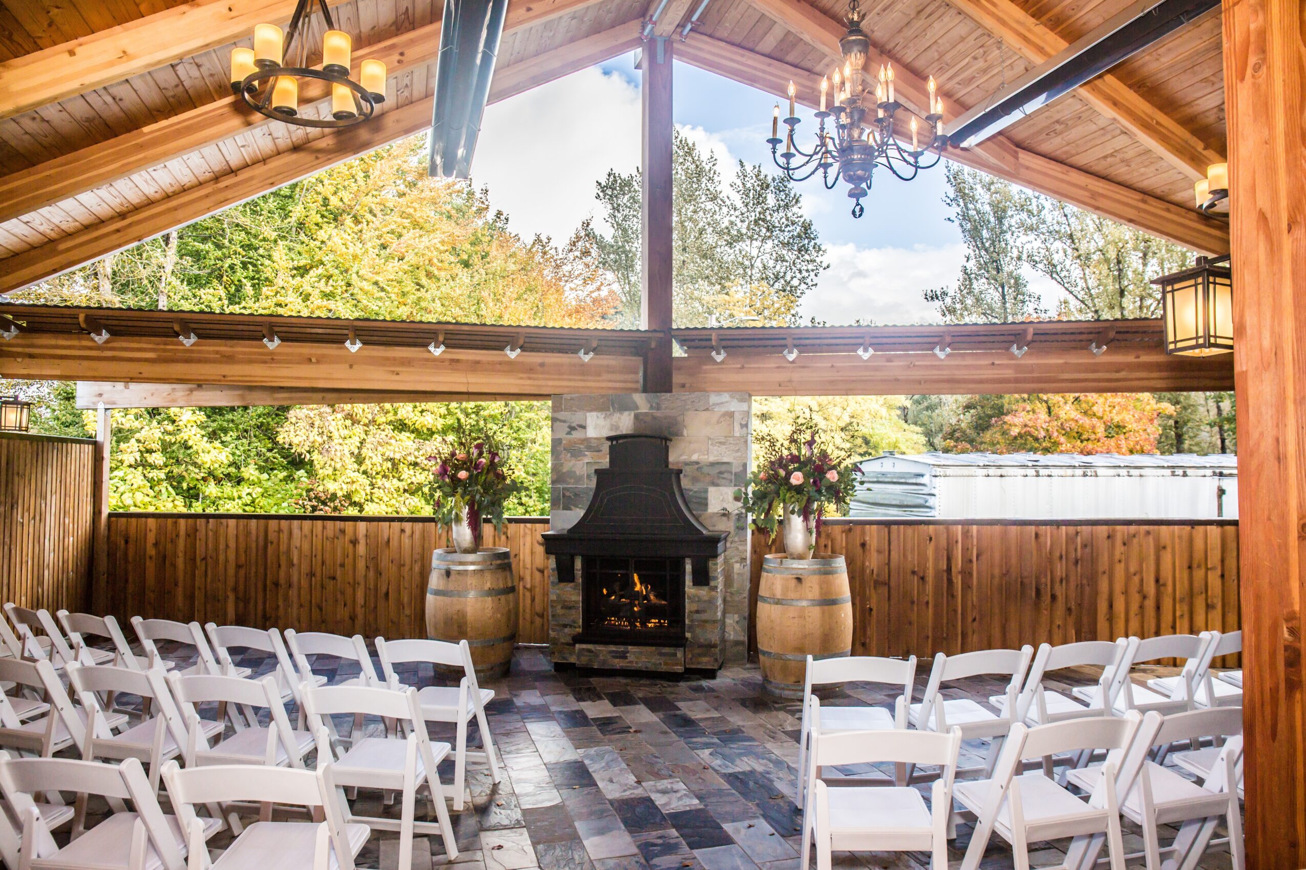 Indoor space with a fireplace, wooden barrels, and white chairs, overlooking trees outside through large windows.