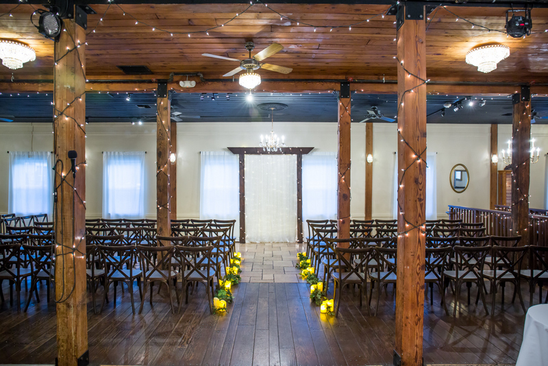 Event hall with wooden beams, chairs arranged in rows, and yellow flowers along the aisle, illuminated by ceiling lights.