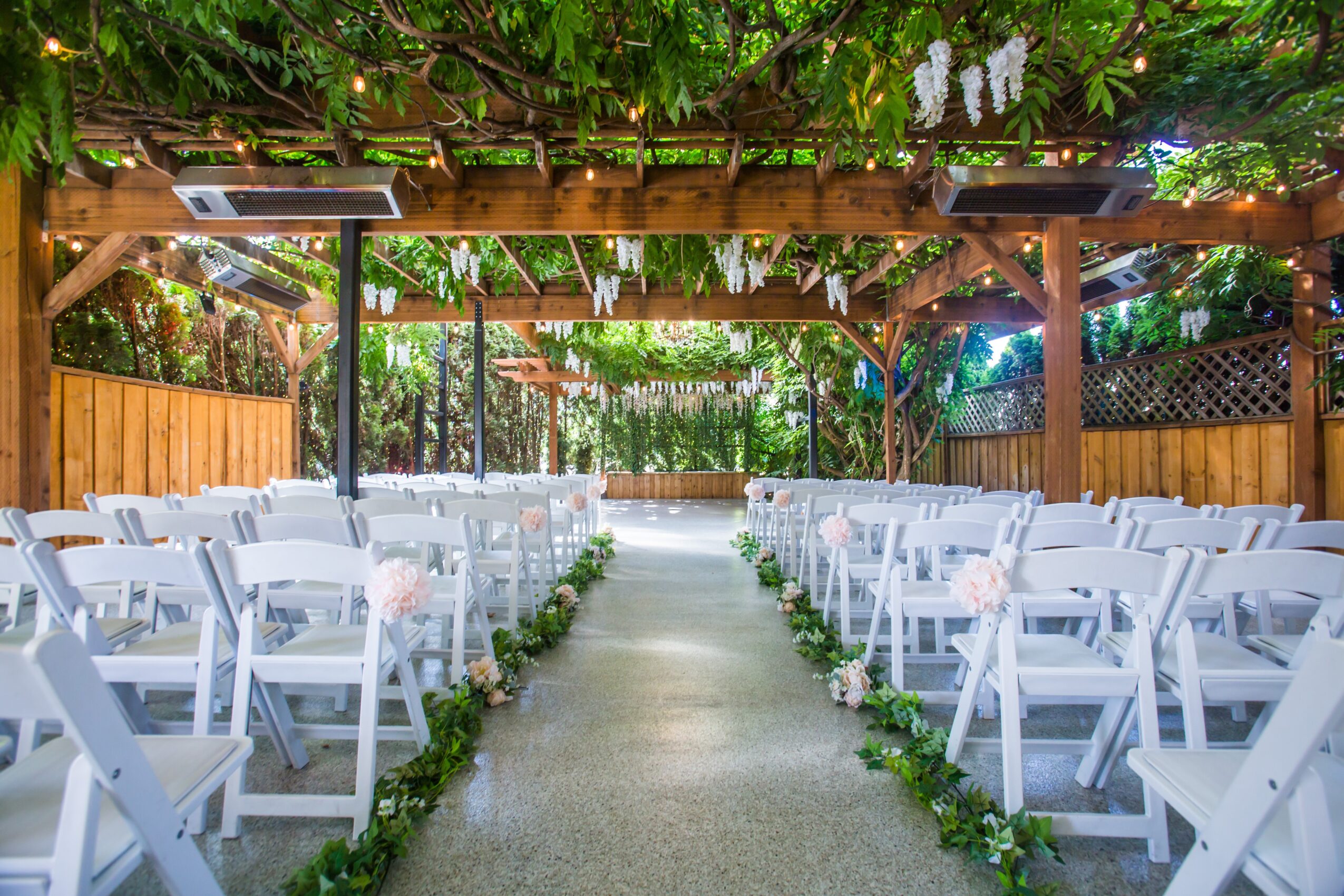 Outdoor wedding ceremony setup with white chairs on either side of a central aisle, decorated with greenery, under a wooden pergola.