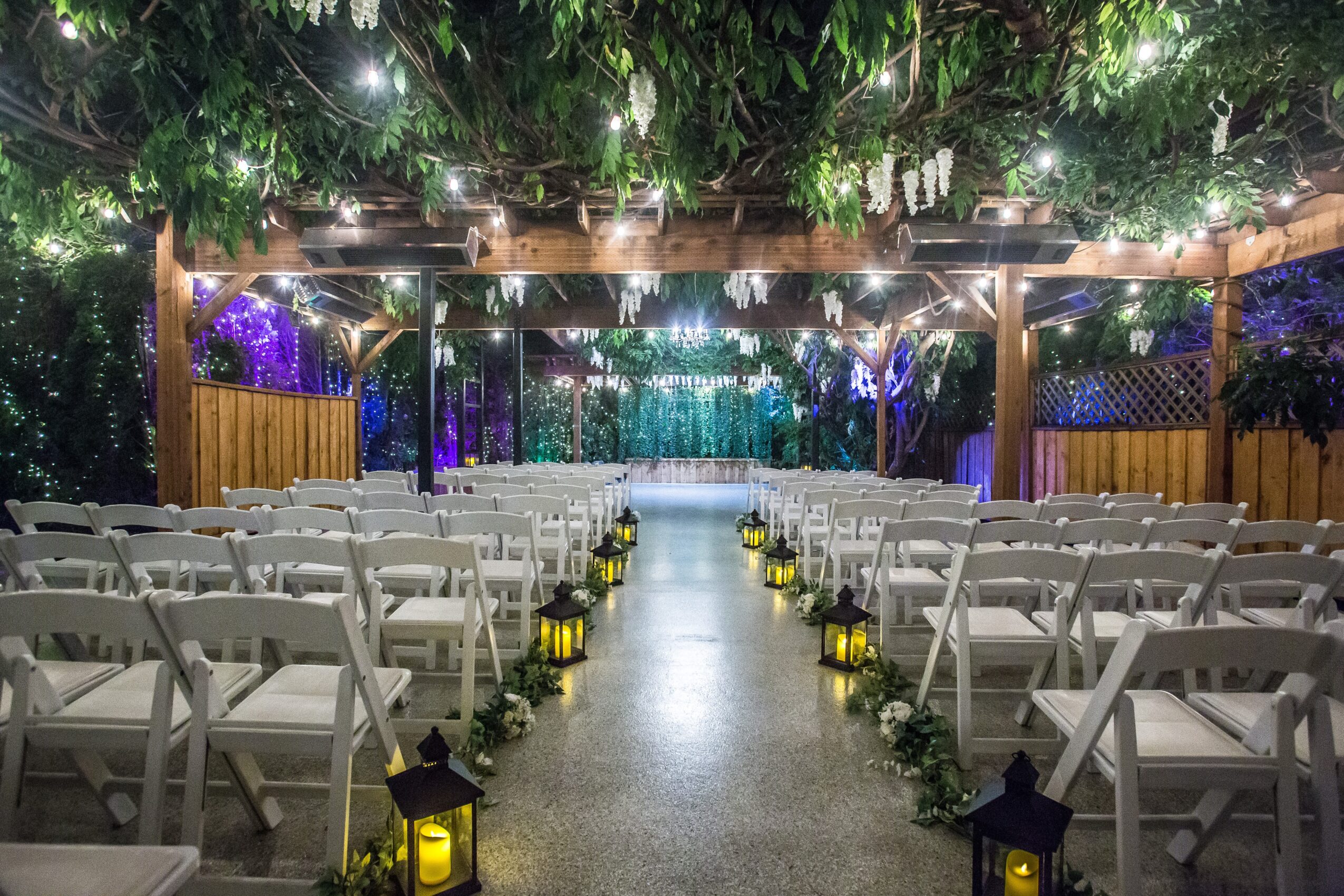 Outdoor wedding ceremony setup with chairs, lanterns, and a decorated stage under trees with string lights.