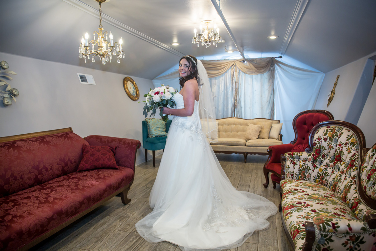 Woman in wedding dress holding bouquet in a decorated living room with vintage furniture and chandeliers.