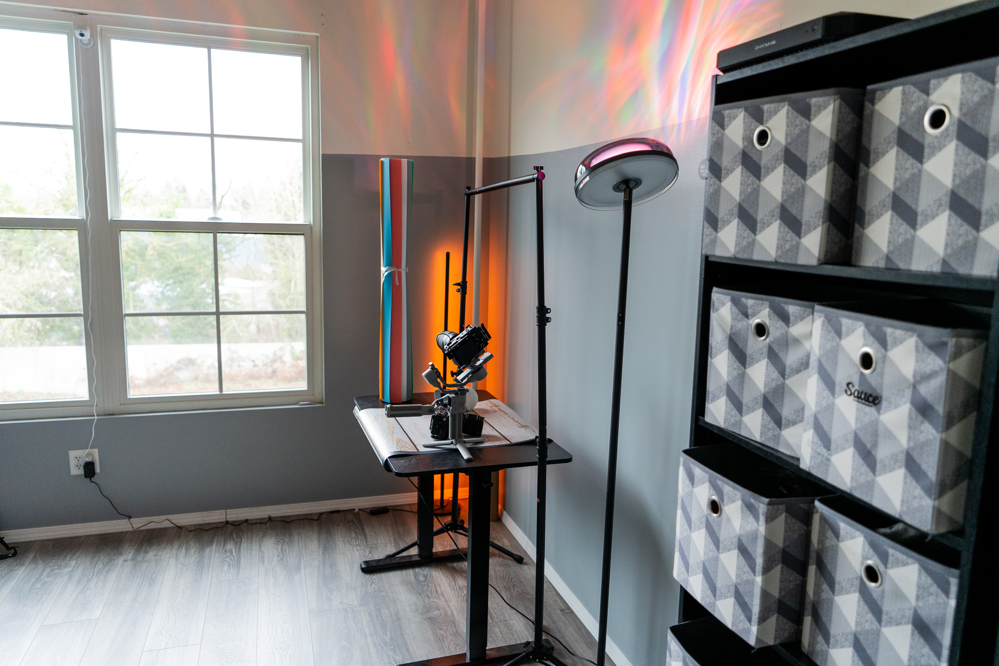 Room with window, microscope on table, and storage boxes on shelves, illuminated by natural light and colorful reflections on wall.