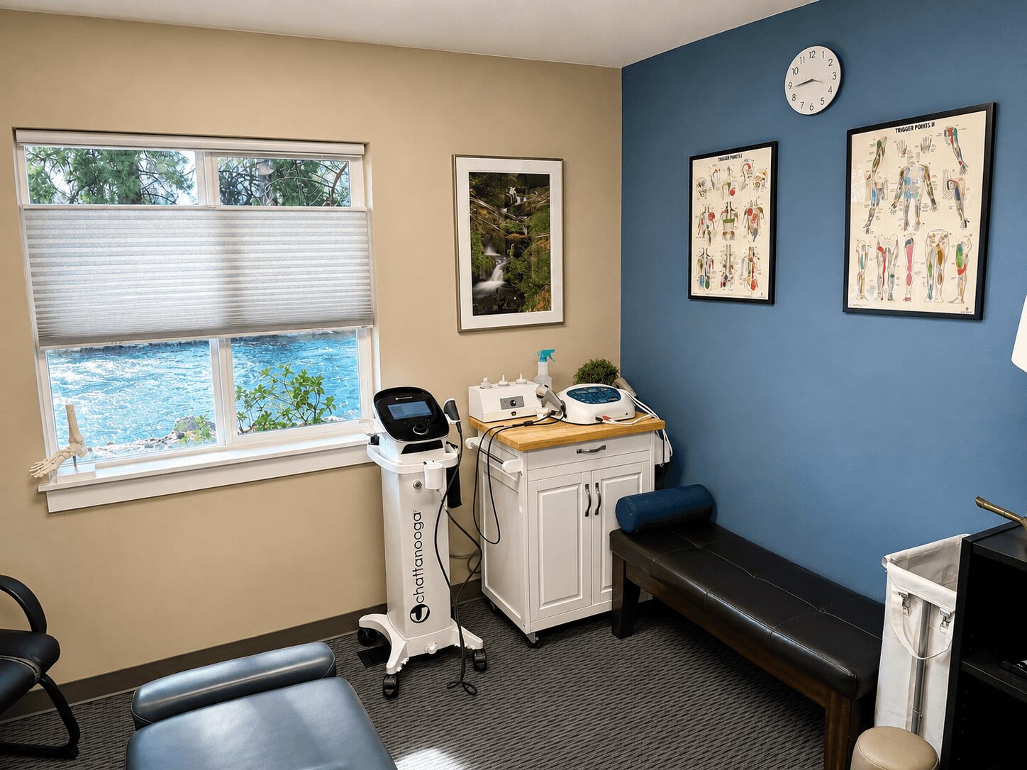 Medical examination room with beige and blue walls, window with blinds, medical equipment, and framed artwork.