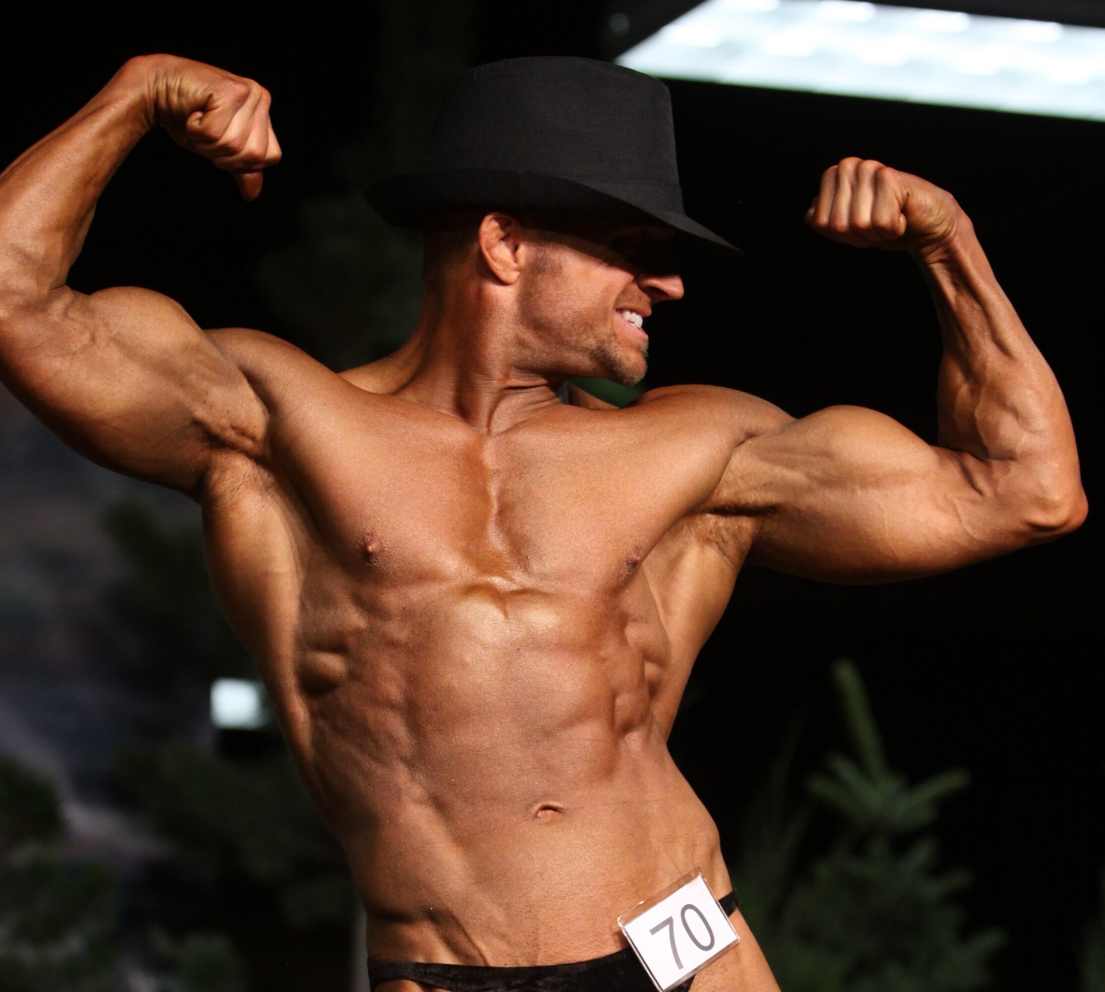 Male bodybuilder wearing a hat, flexing muscles, with a number 70 on waist, in a competition setting.