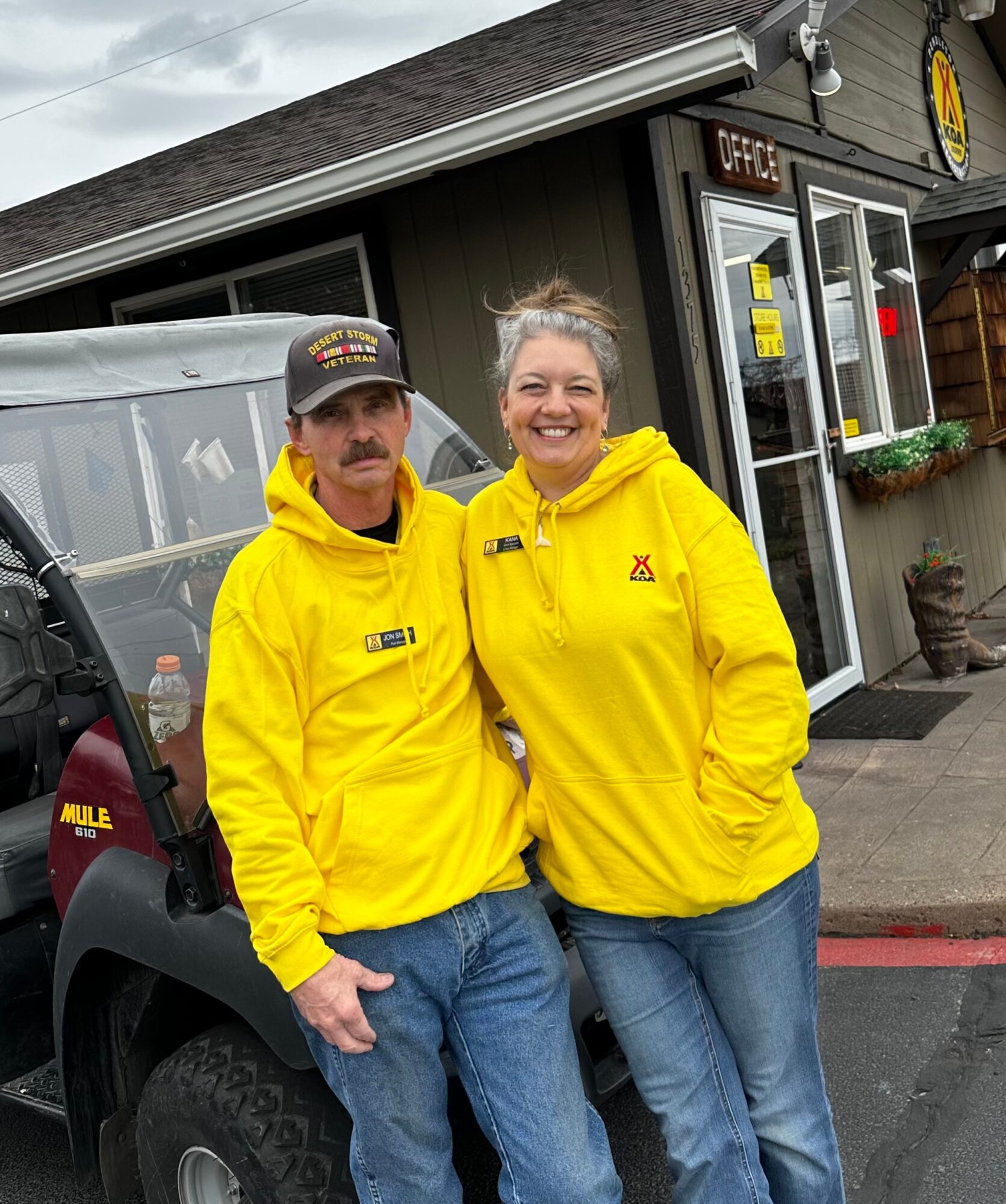 Two people in yellow jackets standing outside a building, smiling, with a vehicle nearby.