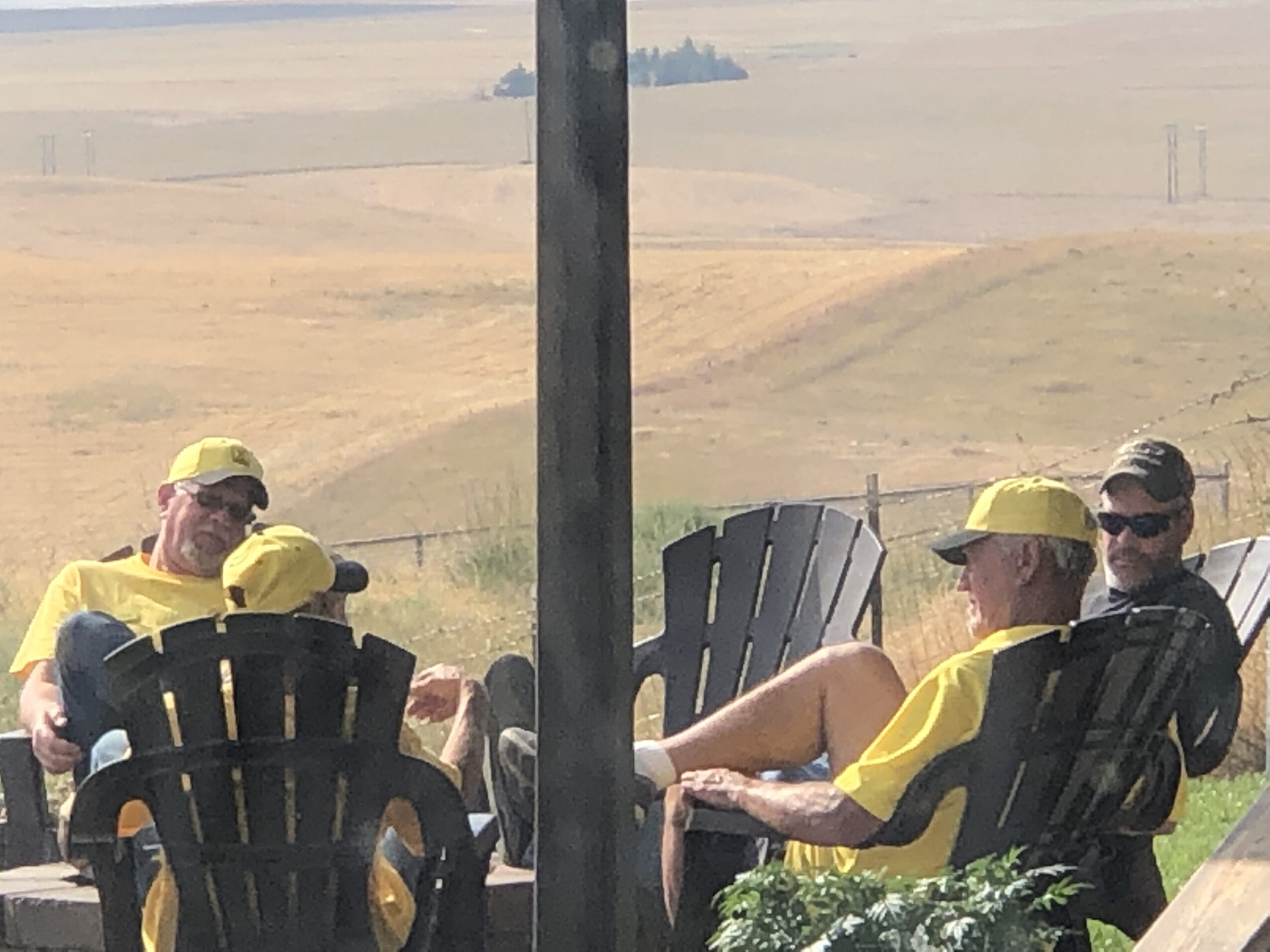Group of people sitting outdoors on black chairs, wearing yellow hats and shirts, with a rural landscape background.
