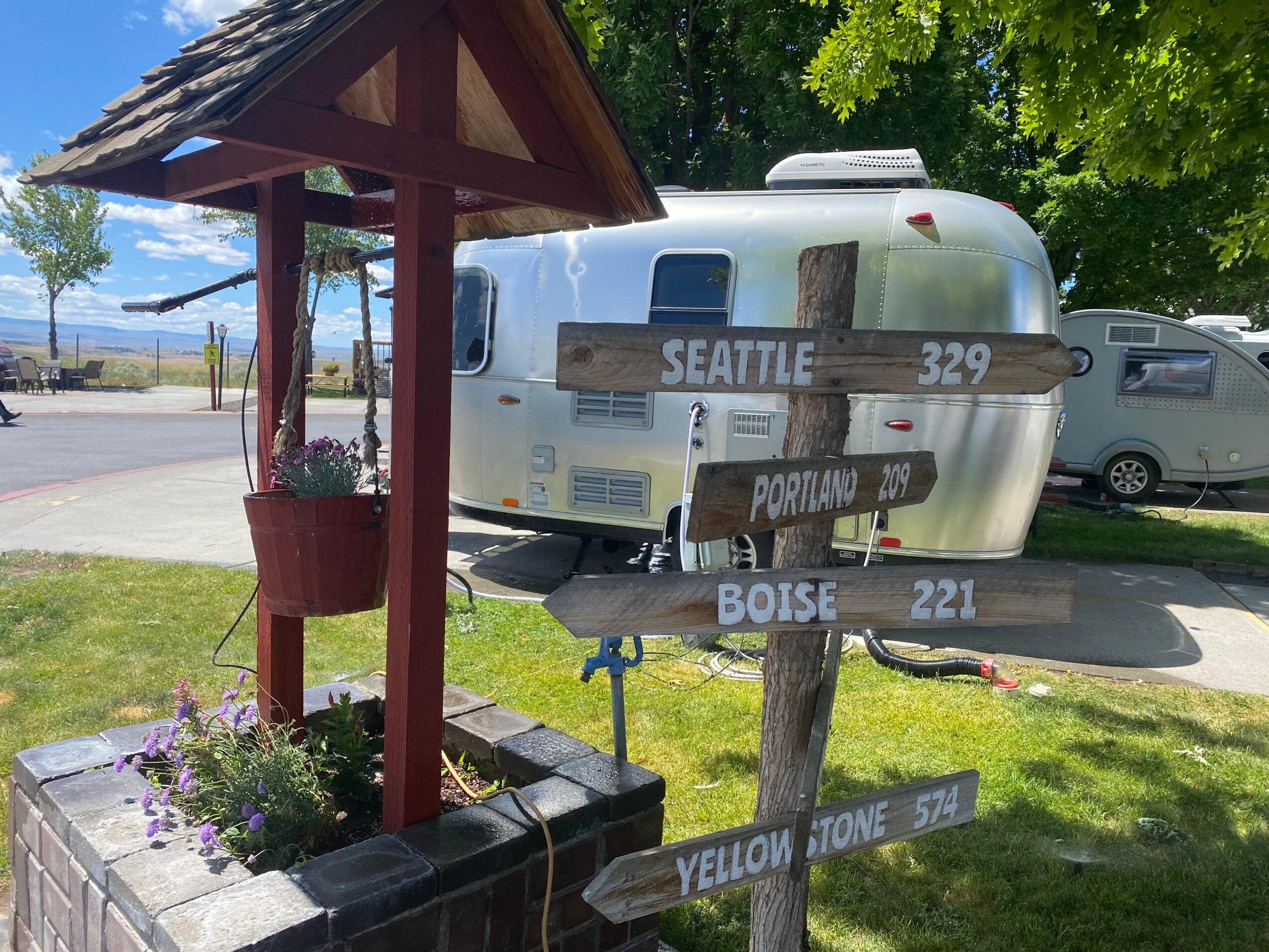 A signpost with four directional signs in front of a shiny silver camper, with trees and parked cars in the background.