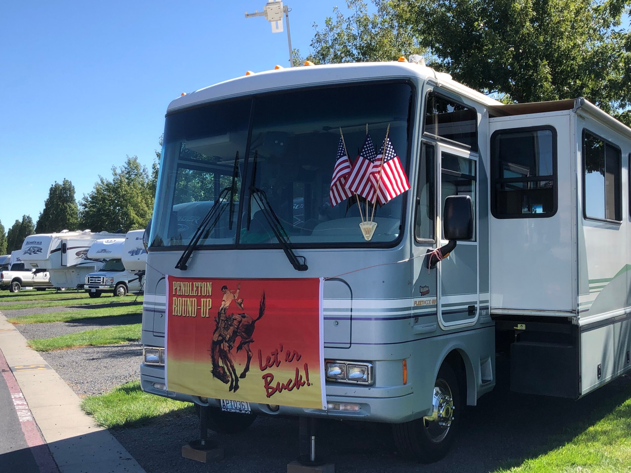 RV parked outdoors with American flags and a sign showing a horse rider and text, under a clear blue sky.