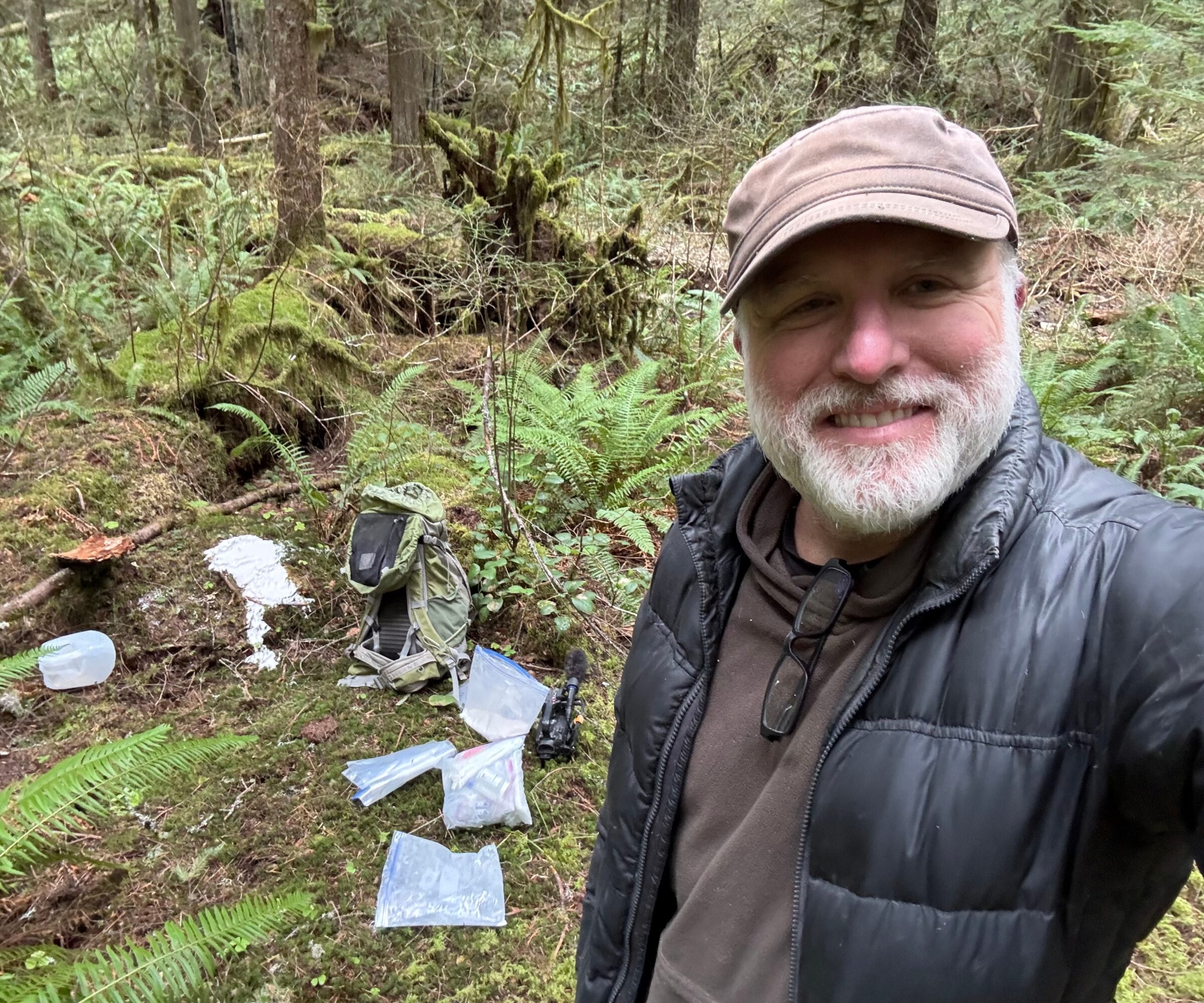 Man with a beard and cap smiling in a forest, with equipment and maps on the ground nearby.