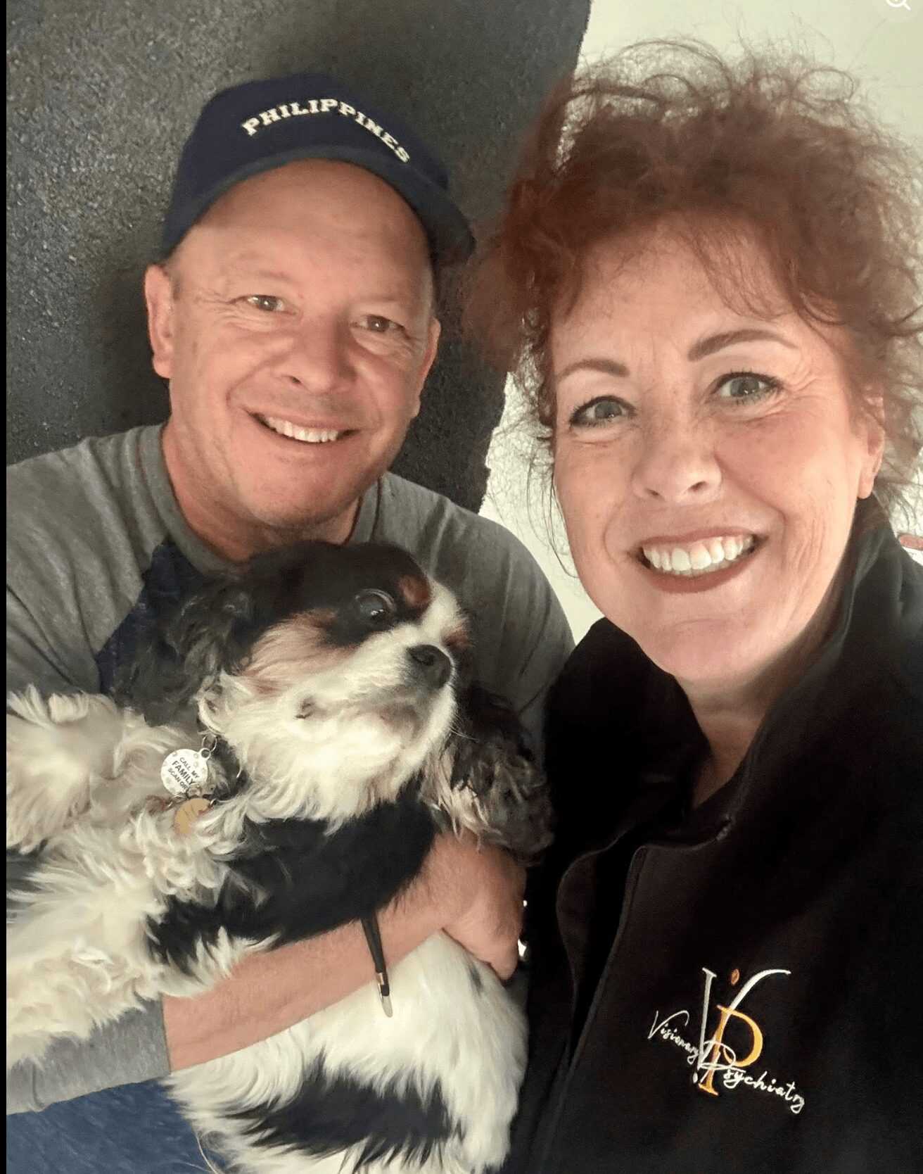 Two smiling people holding a small dog, close together indoors, with a plain background.