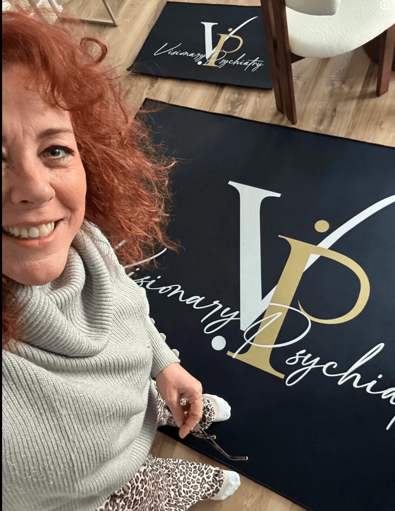 Woman with curly red hair smiling, wearing a light gray turtleneck, holding a phone, next to a black sign with white and gold lettering.