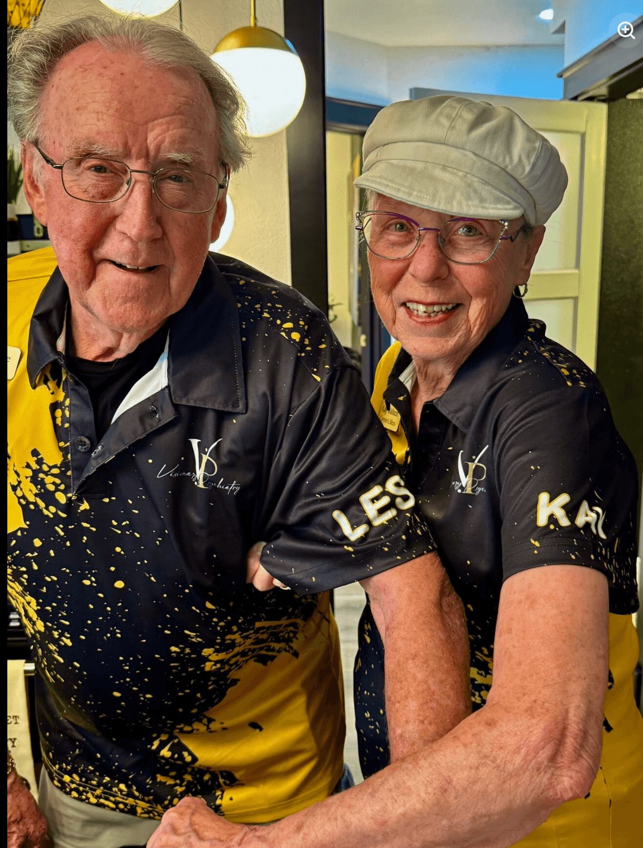 Two elderly people smiling, wearing matching black and yellow shirts, in a room with warm lighting and a yellow cabinet.