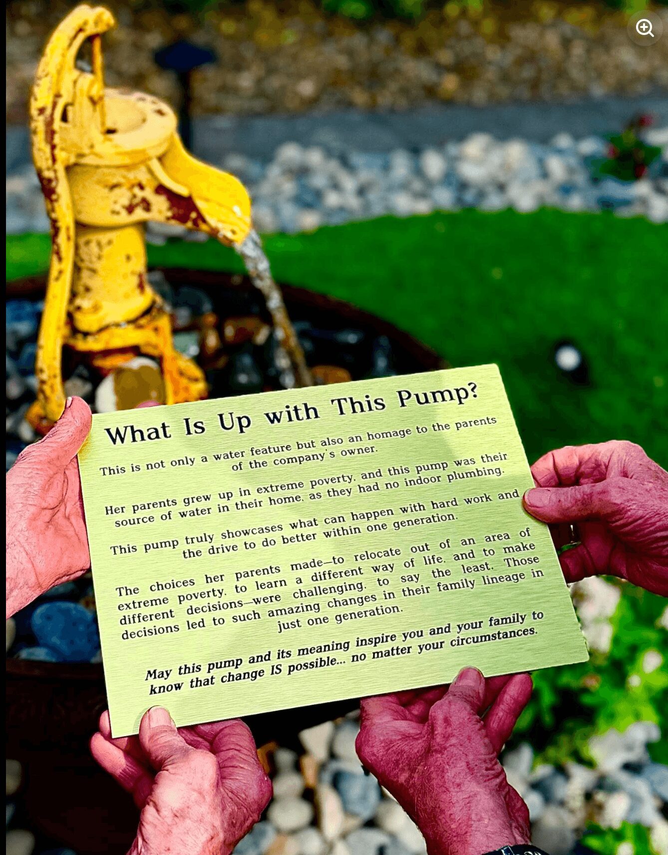 Person holding a yellow fire hydrant-shaped water pump with a card explaining its function outdoors, green grass and rocks in background.