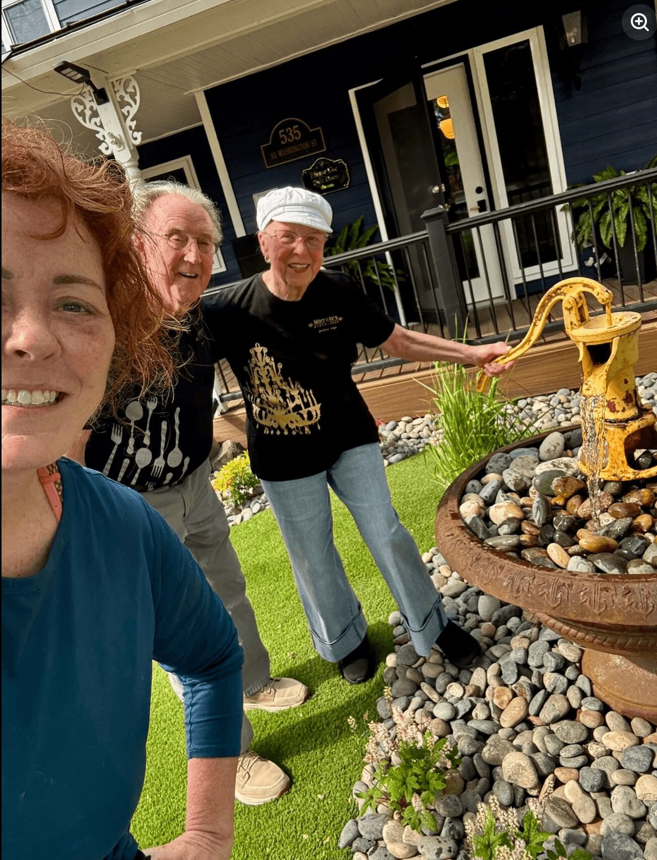 Three people smiling outdoors near a decorative fountain with rocks, plants, and a black house with white trim in the background.