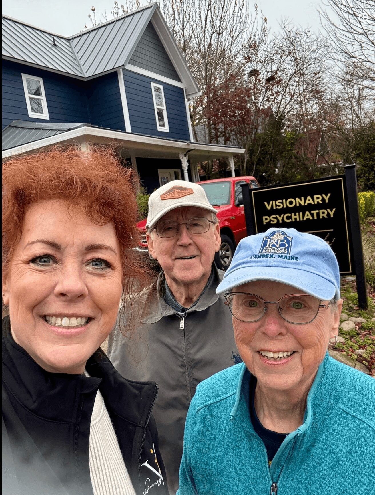Three people smiling outdoors in front of a house and a sign that reads 'Visionary Psychiatry.'