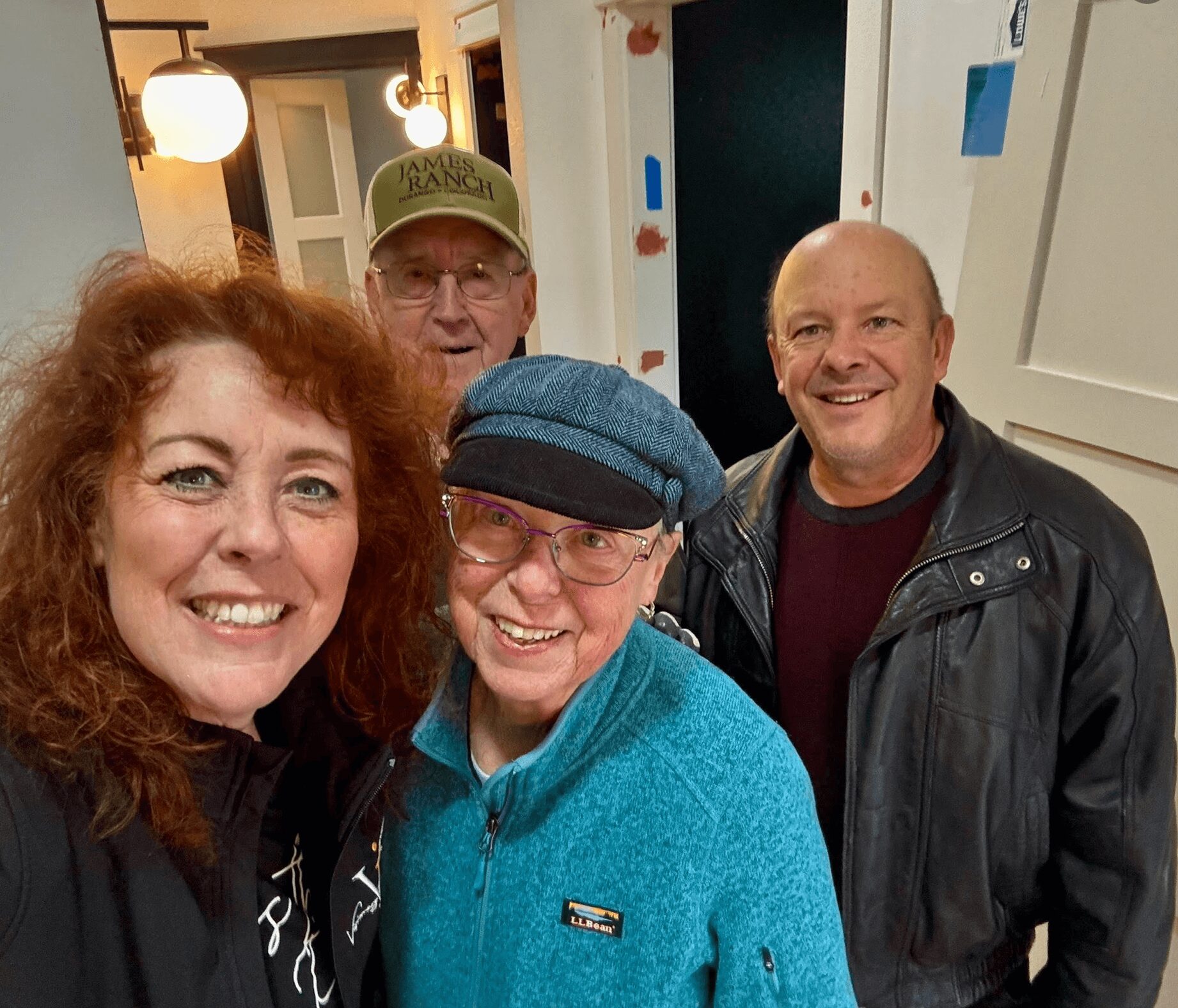 Four people smiling indoors, two women and two men, with warm lighting and a door in the background.