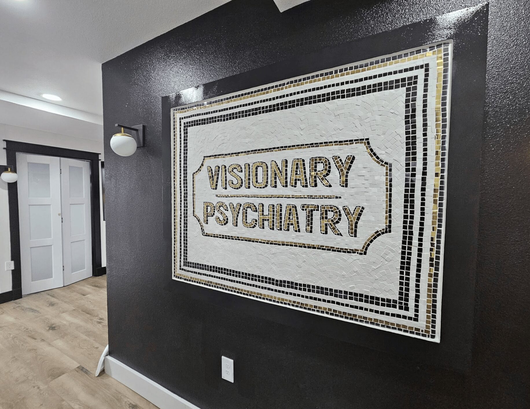 Sign on black wall reads 'Visionary Psychiatry' in mosaic tiles, with a hallway and door visible nearby.