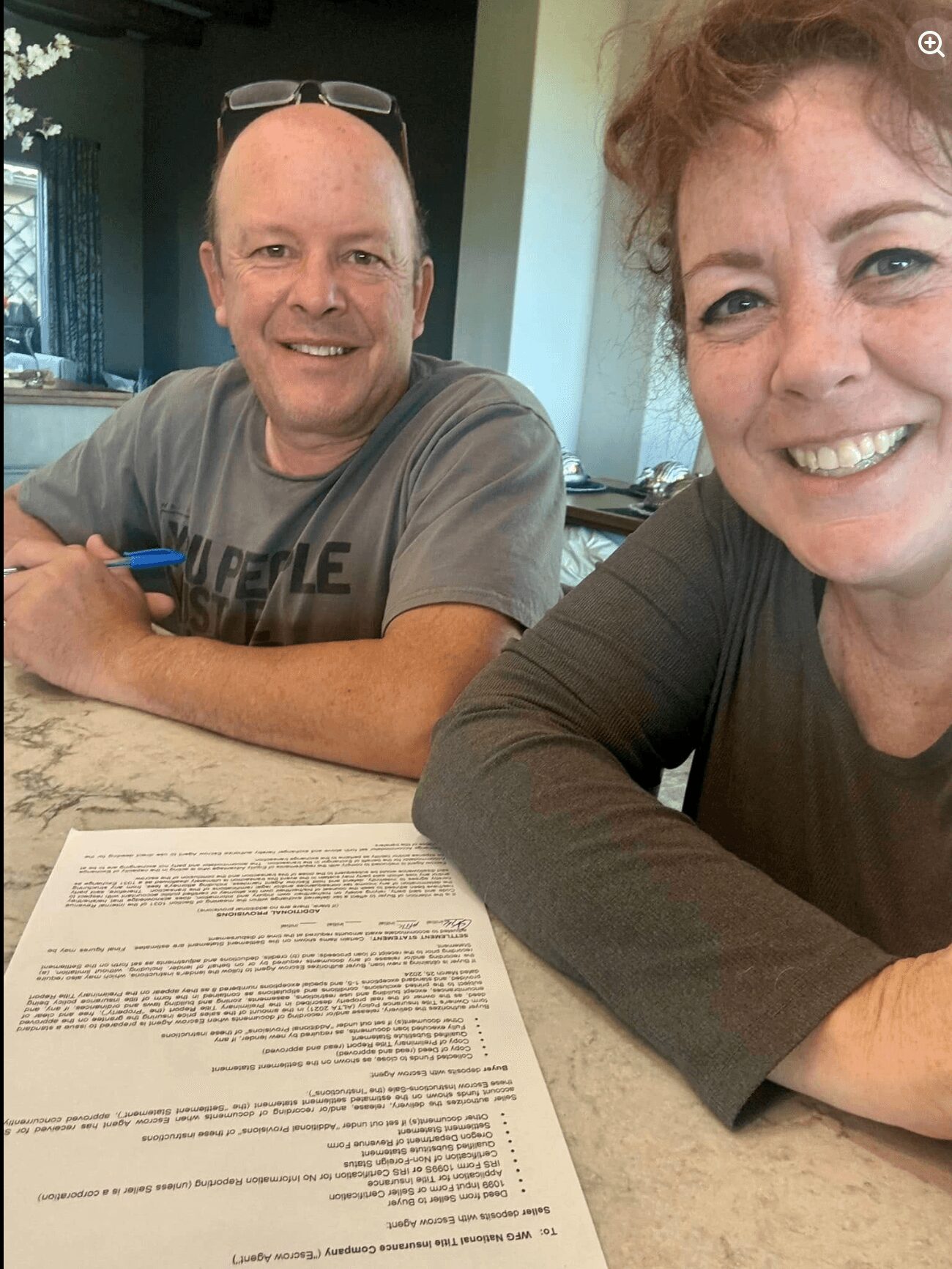 Two people smiling at a table with a document, one with glasses on head, in a room with natural light.