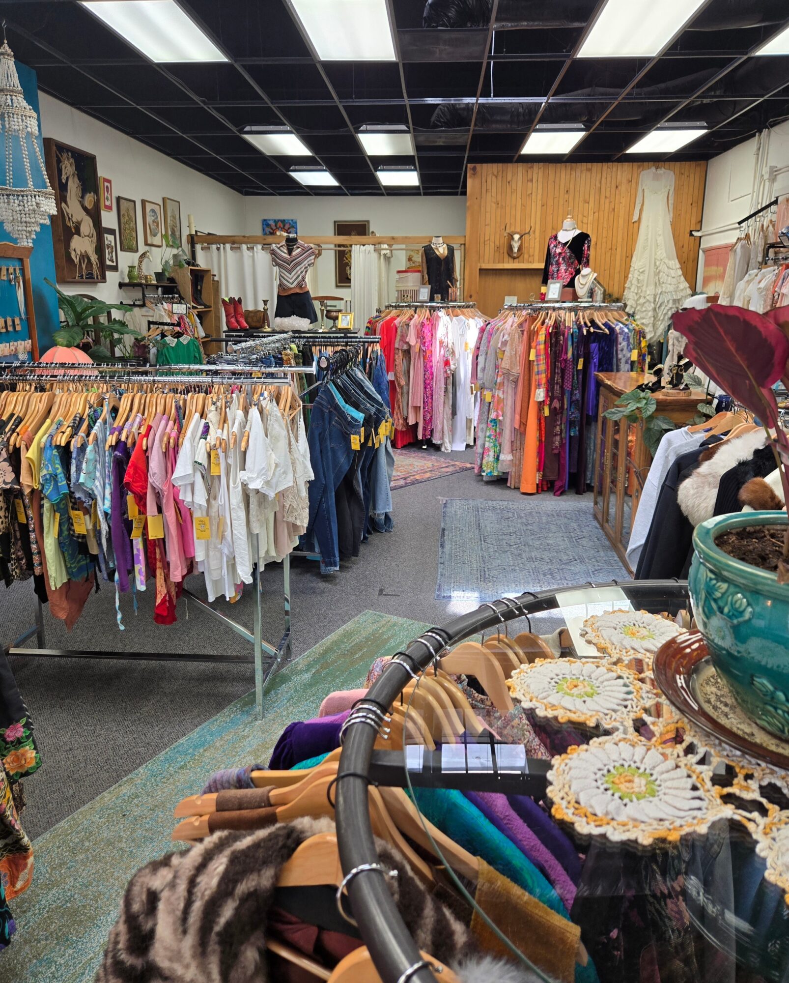 Clothing store interior with racks of colorful clothes, wooden walls, and display tables, under a black ceiling with fluorescent lights.