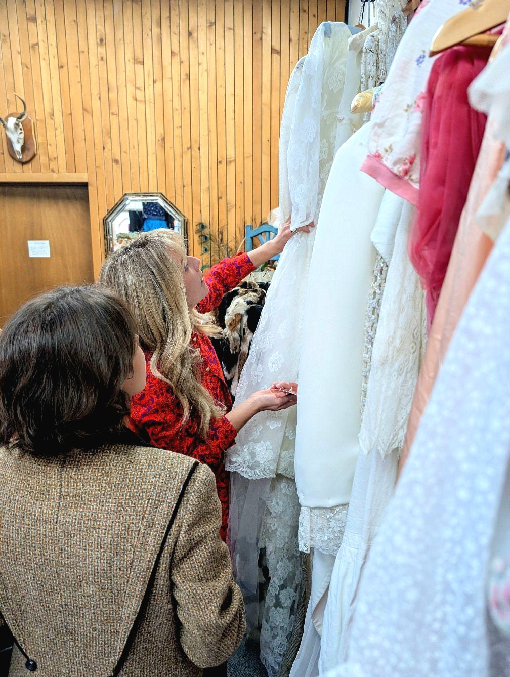 Children browsing white and pink dresses on a clothing rack in a store with wooden walls.