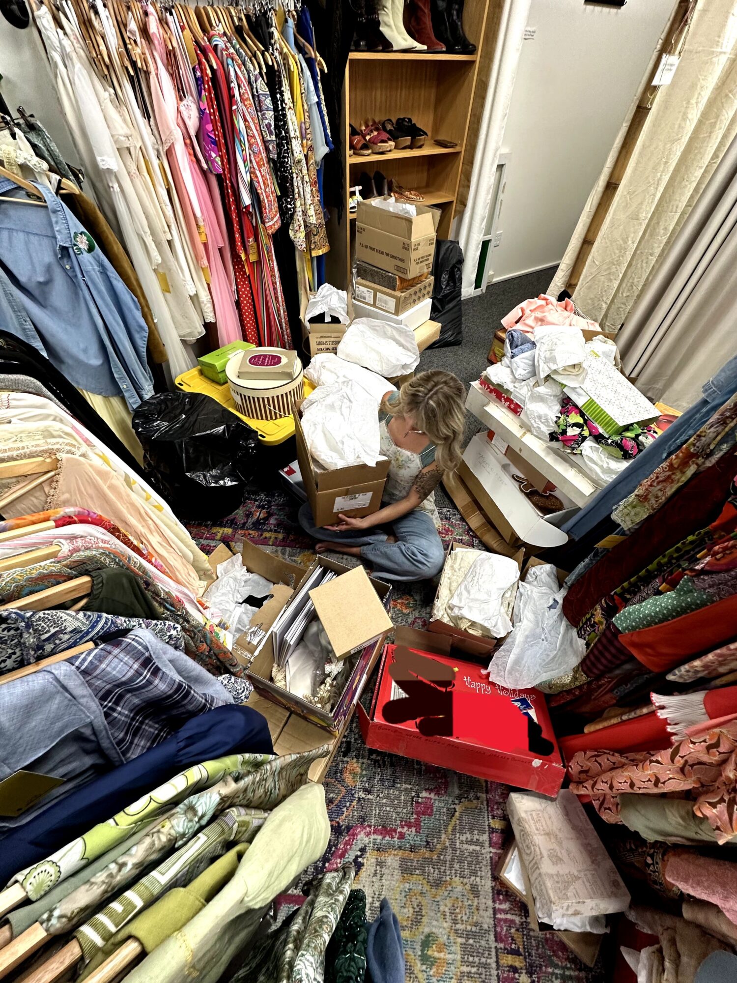 Child sitting on the floor surrounded by clothing, boxes, and packaging in a cluttered room.