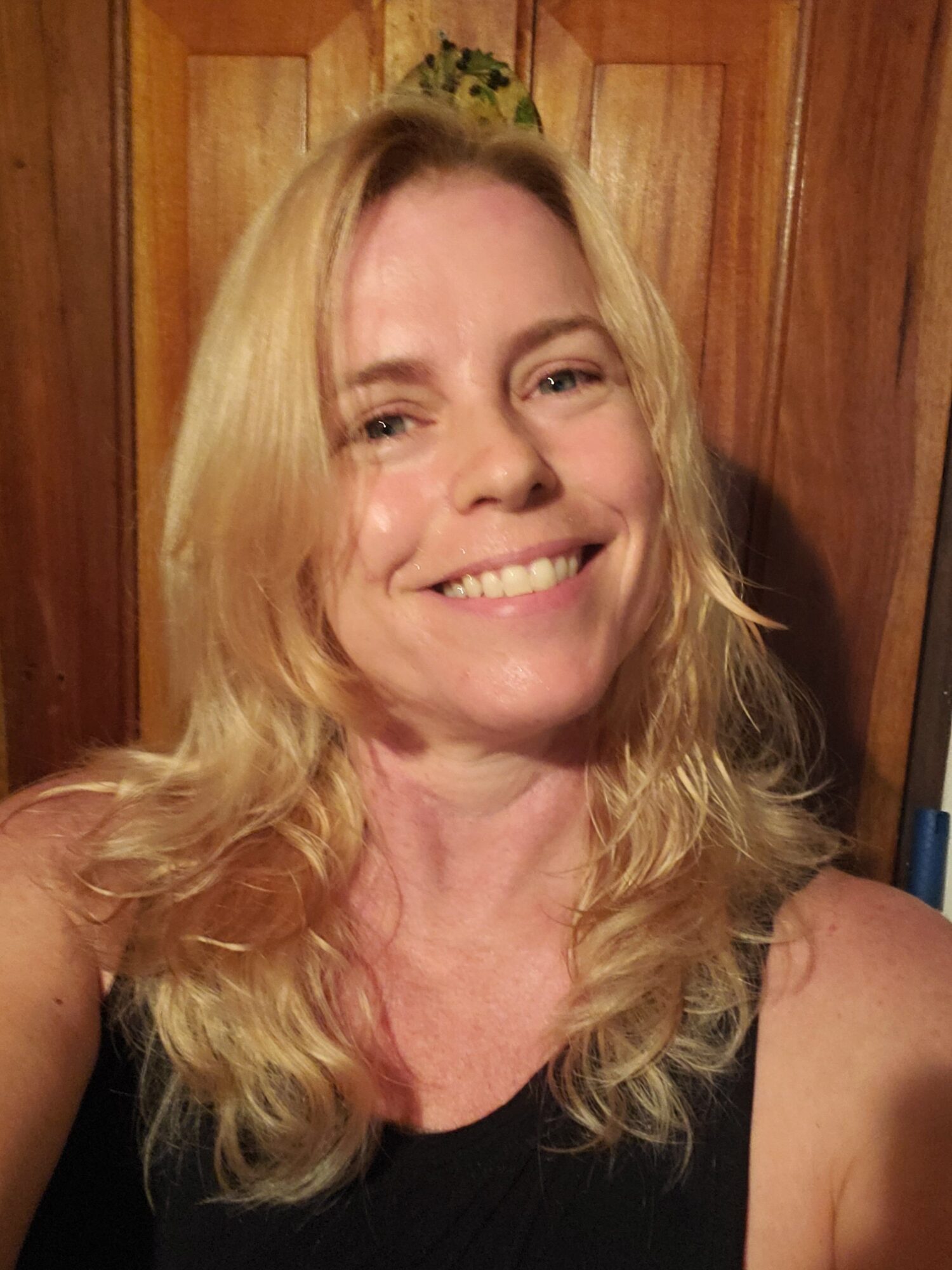 Woman with long blonde hair smiling, wearing a black top, standing in front of wooden background.