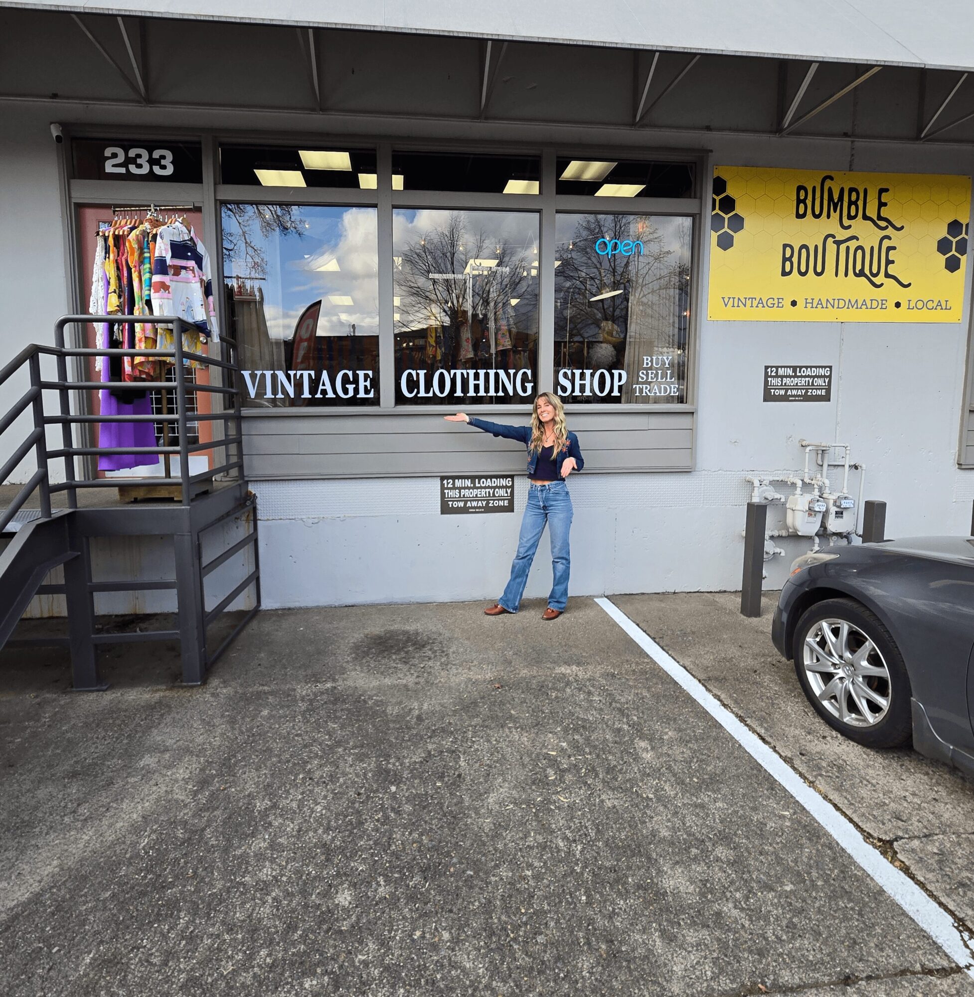 Person standing outside vintage clothing shop with large windows, store sign, and parked car nearby.